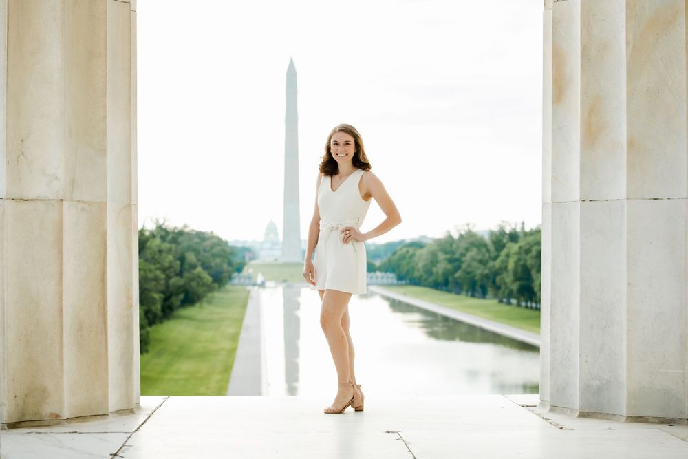 Female in white dress poses between marble columns with Washington Monument and reflecting pool visible in background. Washington DC.