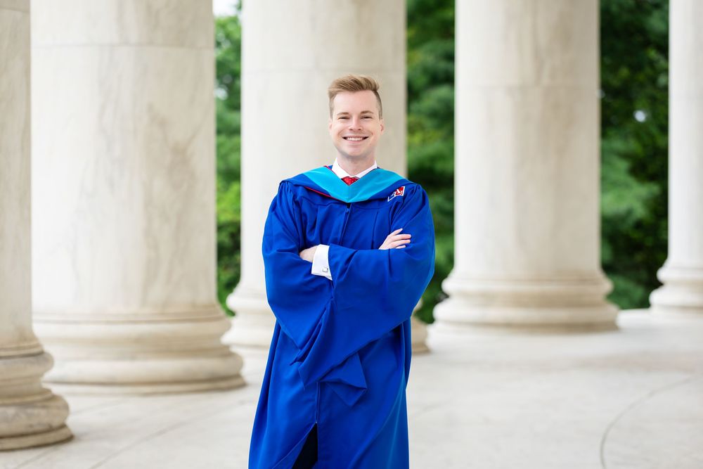 Young man in blue graduation gown and cap stands confidently between white columns at outdoor ceremony.