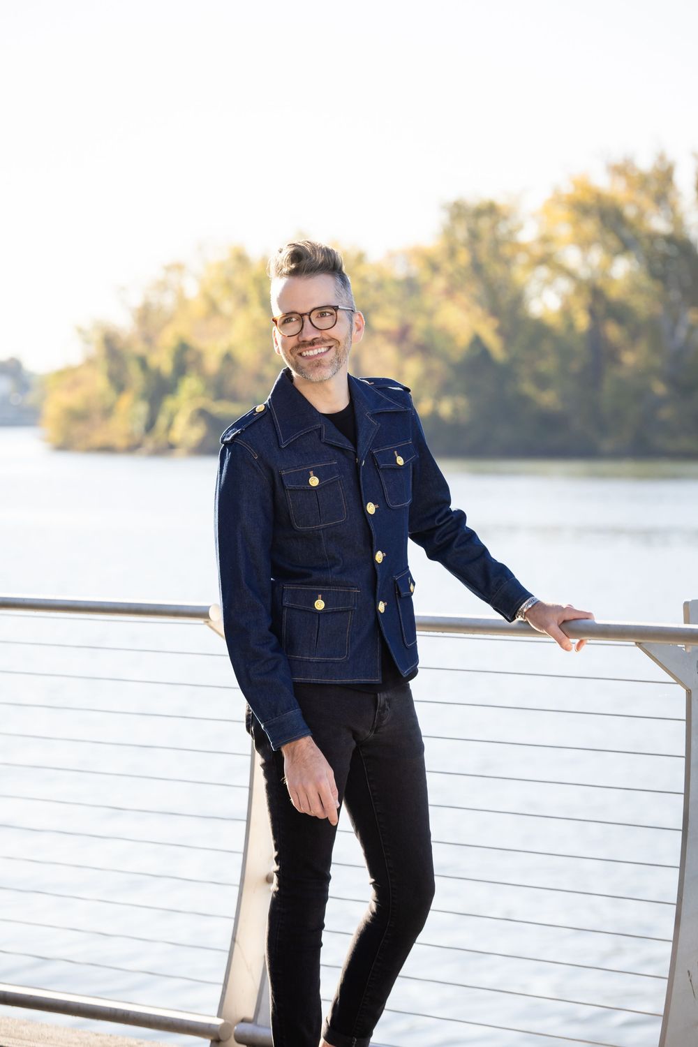 Man in navy denim jacket and black jeans smiling outdoors by waterfront railing on sunny day.