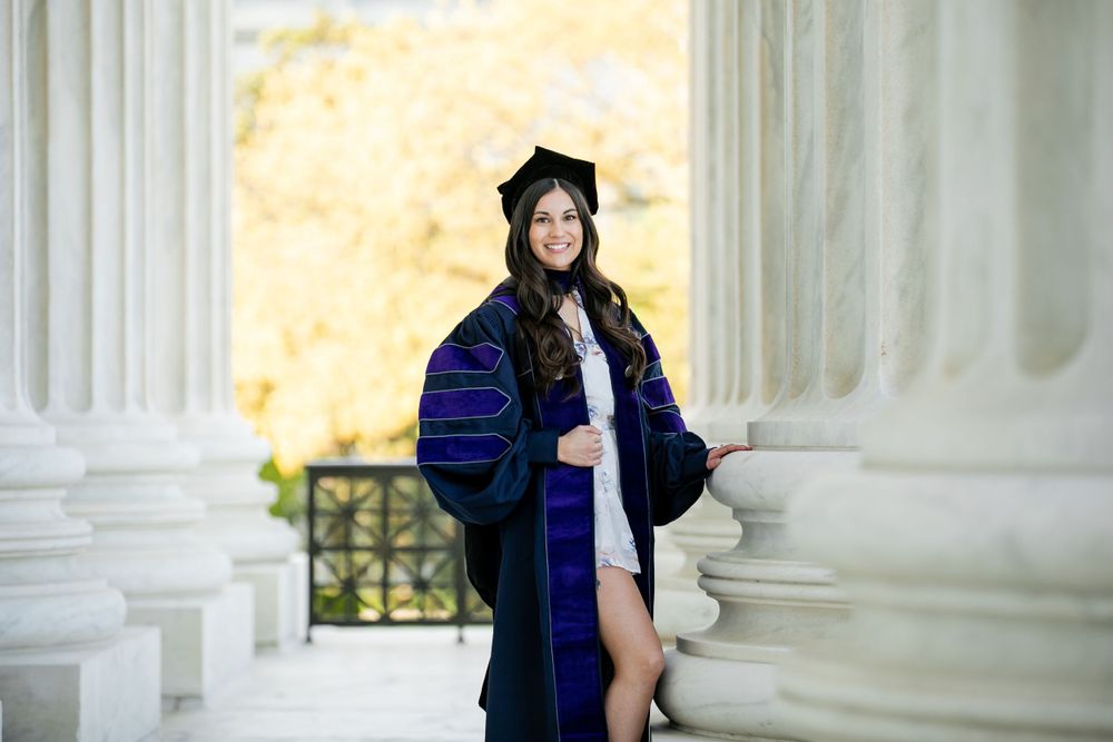 Graduate posing in academic regalia with blue and black graduation gown between white columns outdoors, by Justin Baker Photography.