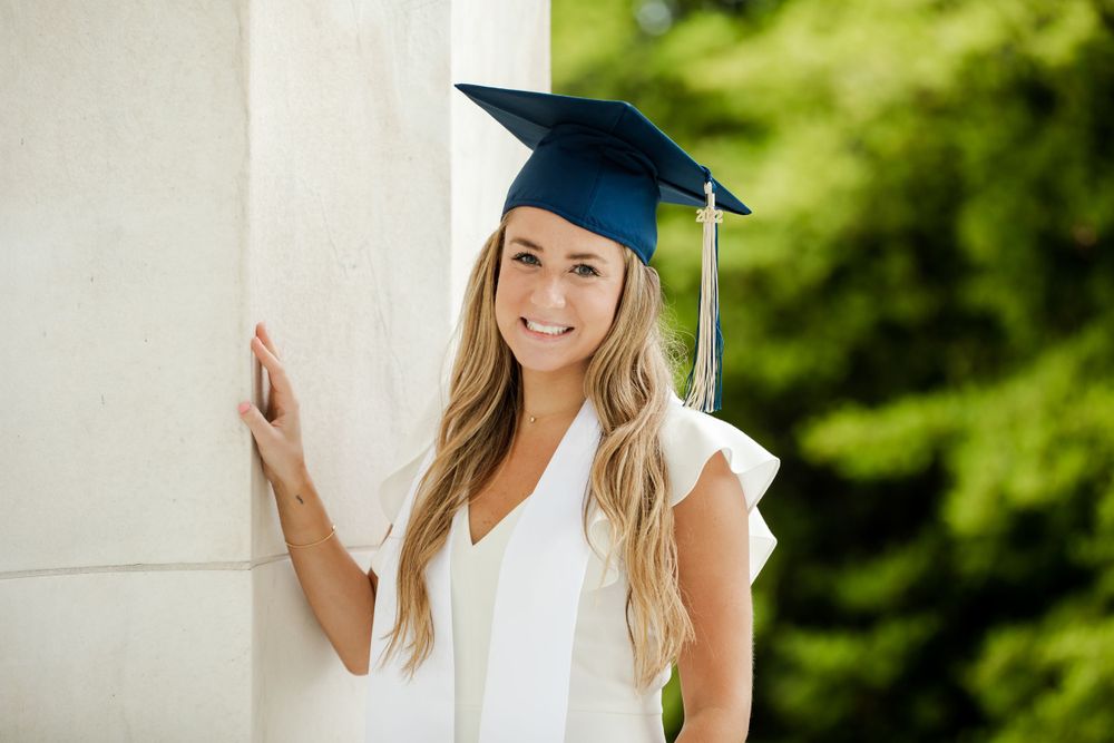 Graduate in white dress and blue cap stands next to white pillar with green trees in background, by Justin Baker Photography. Washington DC.