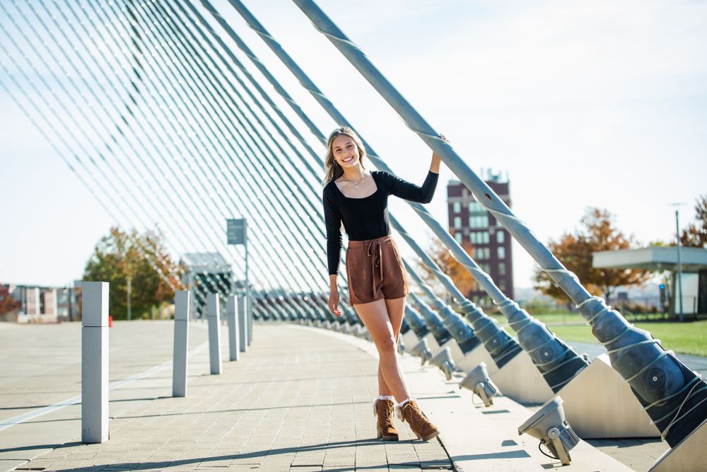 A woman in a black top and brown shorts poses on a modern architectural bridge with diagonal cables on a sunny day in Kansas City.