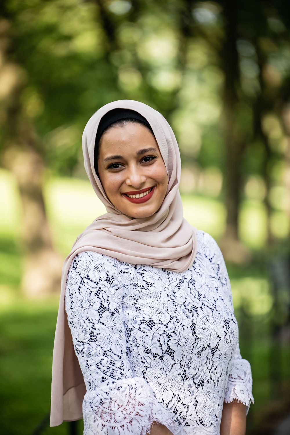 Smiling woman wearing a beige hijab and white lace top poses outdoors in a green park setting.