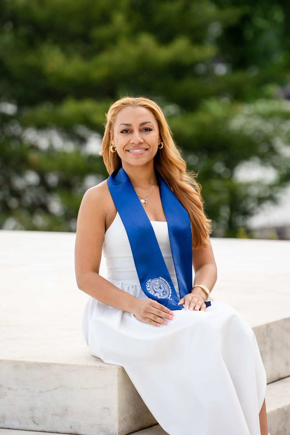 Young woman in white dress with blue graduation stole sits smiling on marble steps outdoors.