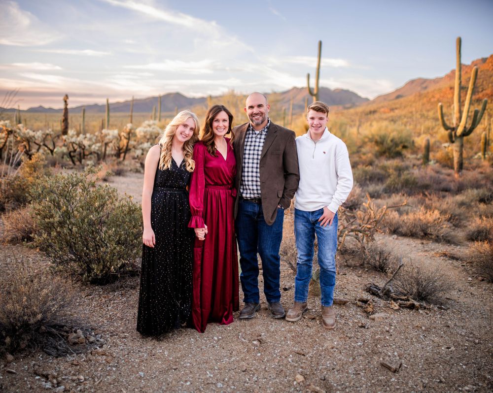 Family of four smiling in a desert setting with saguaro cacti and mountains at dusk.
