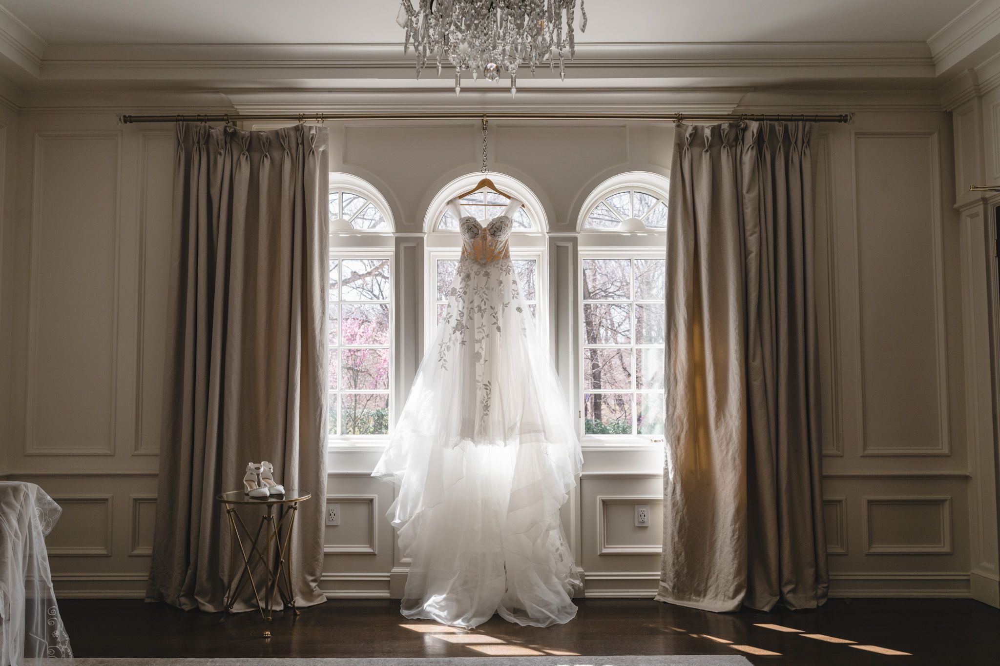 Elegant wedding dress hanging in window with natural light streaming through sheer curtains.