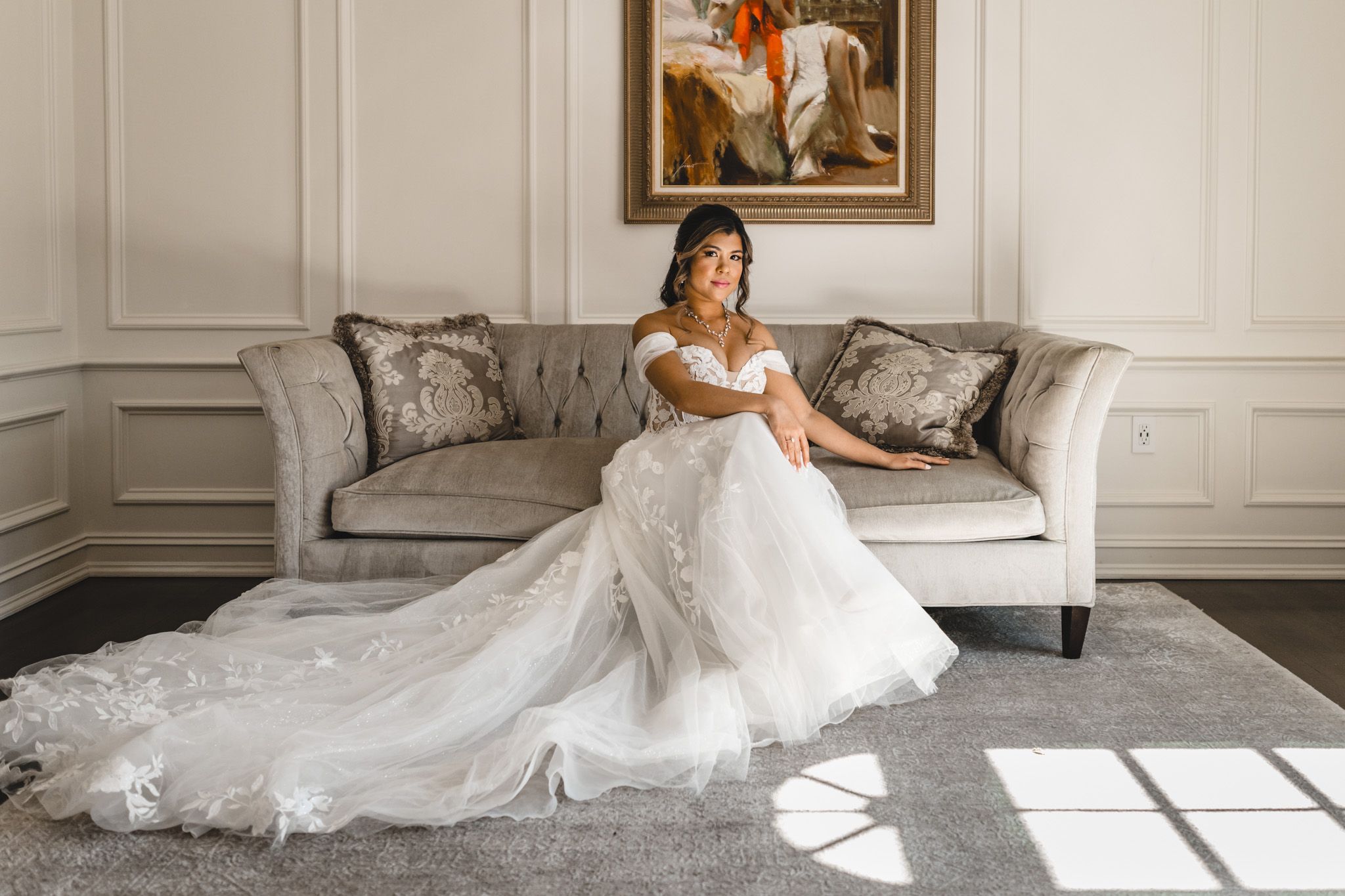 Bride in flowing white gown poses near window in elegant room with classic decor.