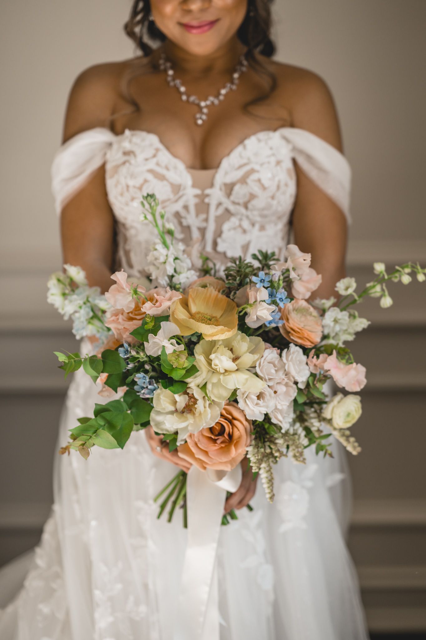 A bride in a white lace gown holds a romantic bouquet of peach and cream flowers with greenery.