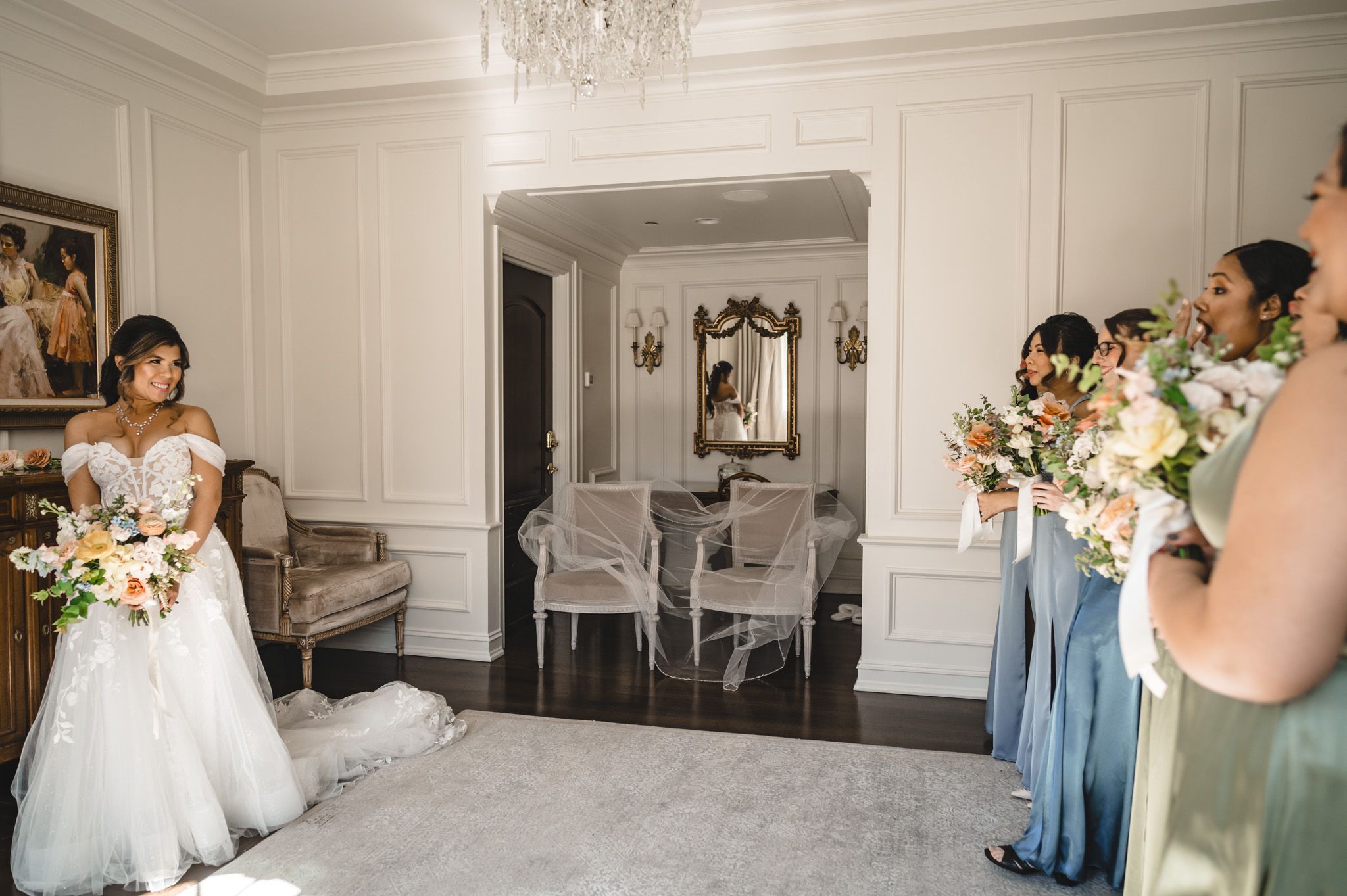 A wedding party gathers in an elegant white room with crystal chandelier and floral bouquets.