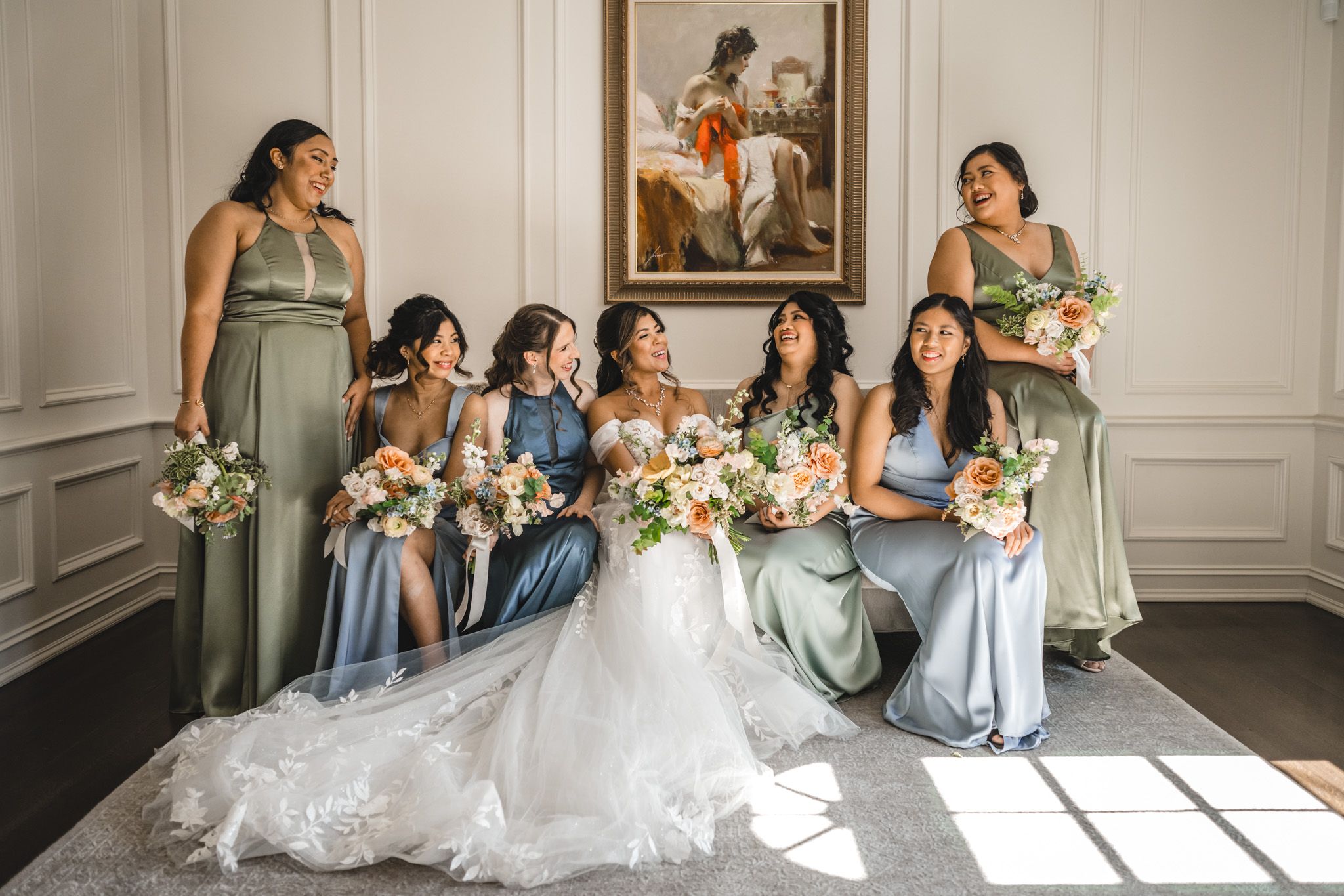 A bridal party poses together in sage and blue dresses holding floral bouquets.