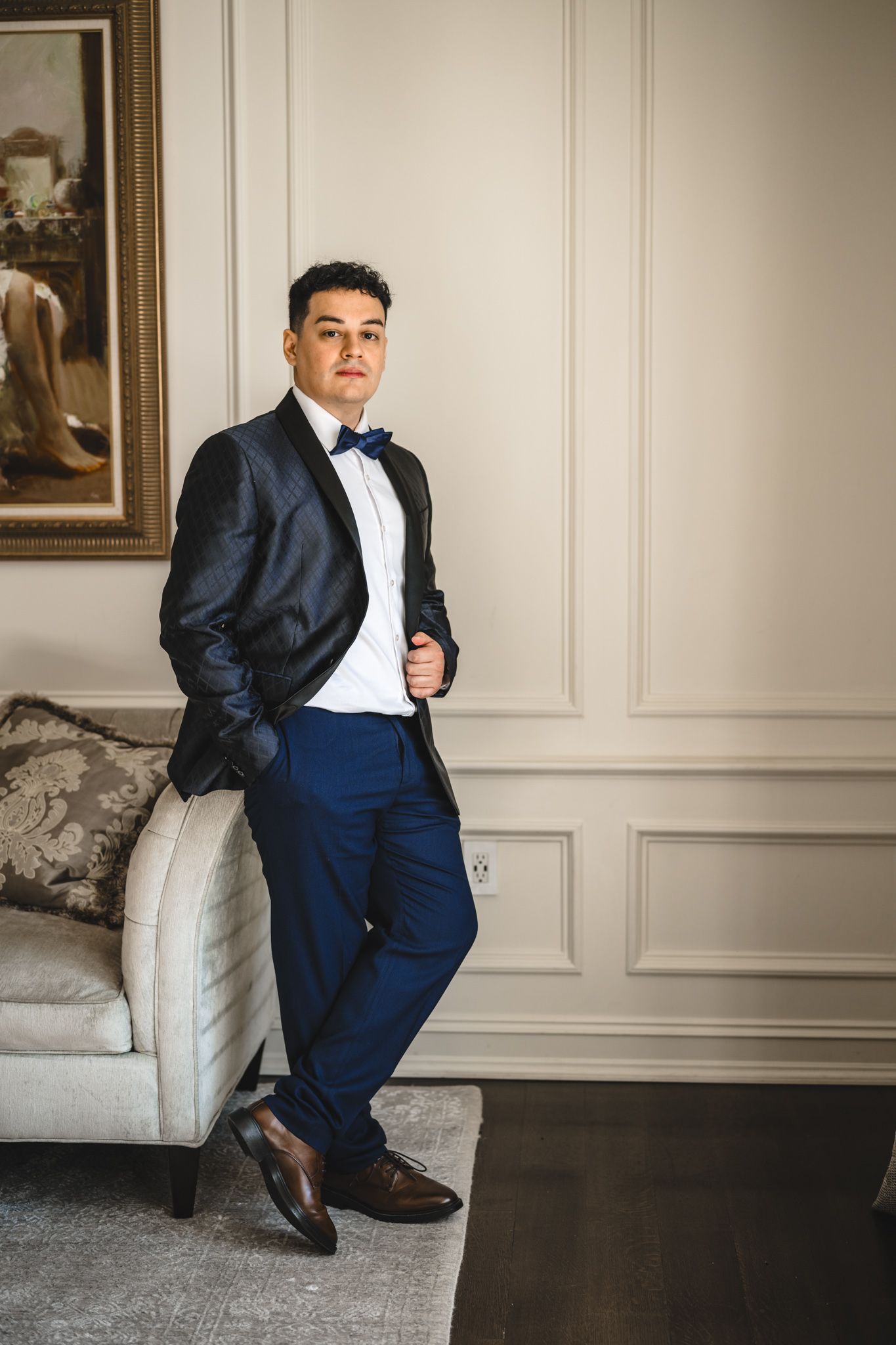 A person in a navy blue tuxedo and bow tie poses elegantly on a white chaise lounge.