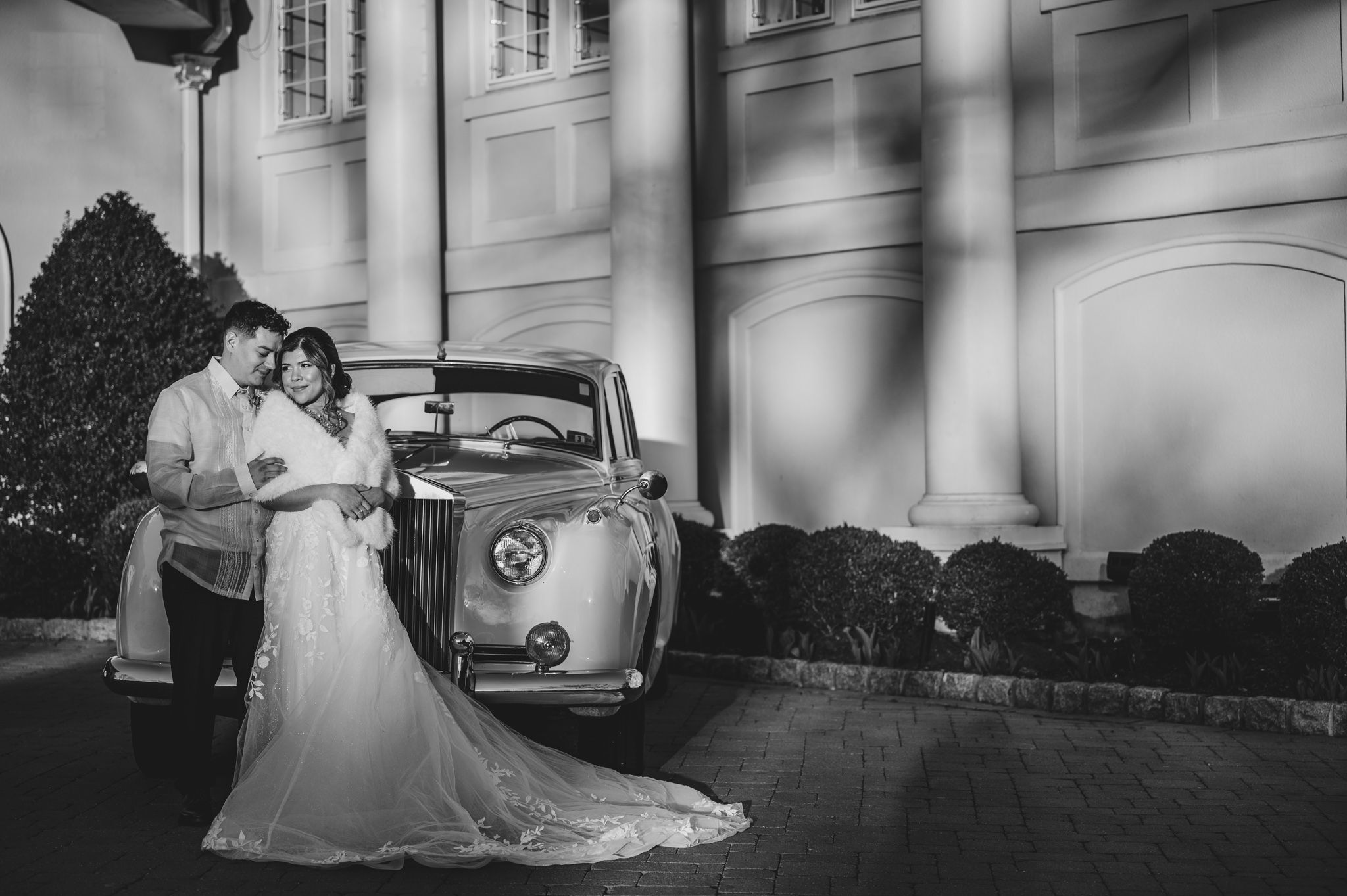 Black and white portrait of couple by classic car with grand architectural columns in background.