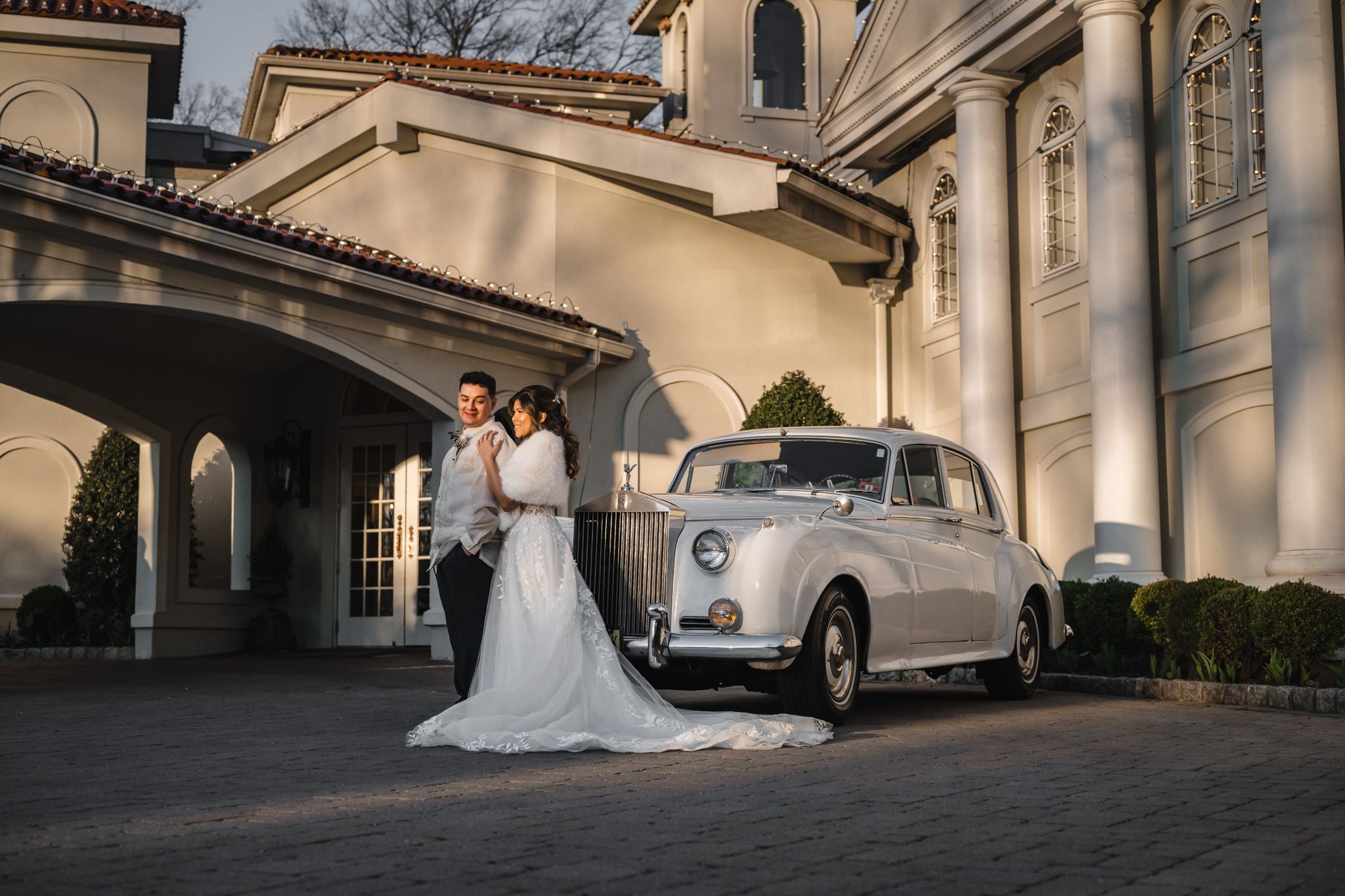Elegant wedding photo with couple posing next to vintage white car outside classic architecture.