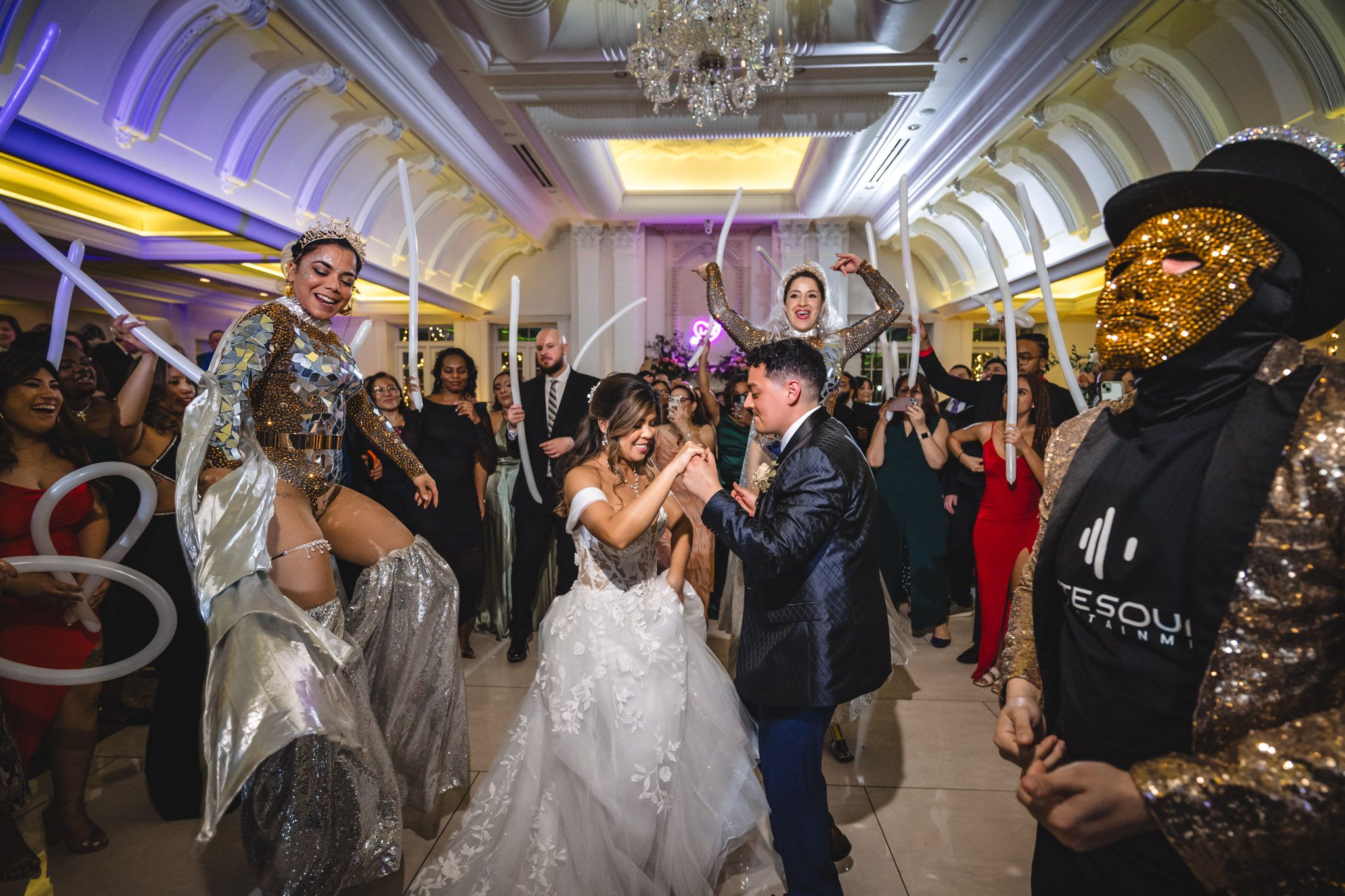 Wedding guests dance and celebrate on a ballroom dance floor illuminated by elegant chandeliers and purple lighting.