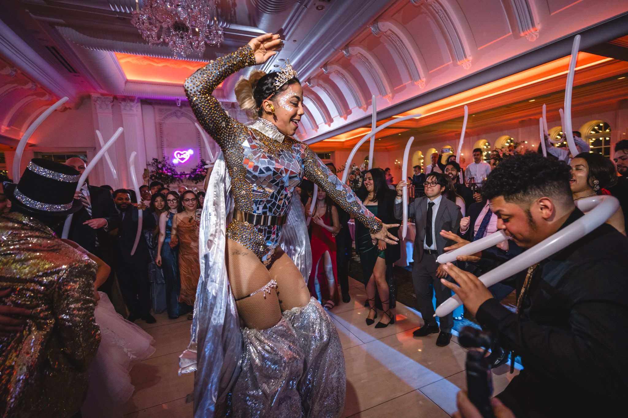 A performer in a silver costume dances energetically at a wedding reception with colorful mood lighting.