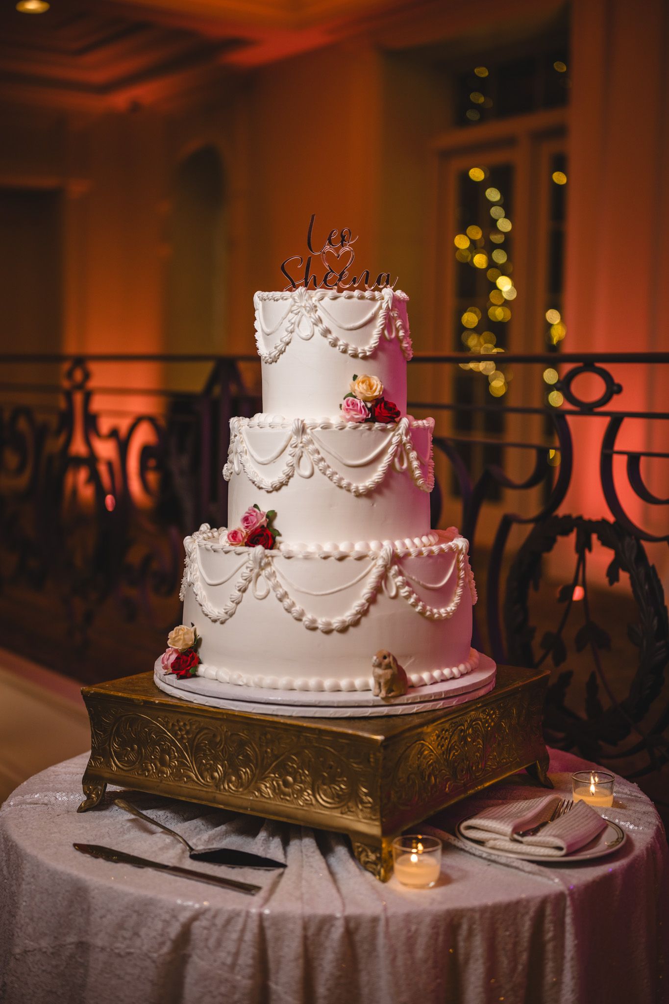Elegant white wedding cake with pearl details and burgundy flowers displayed on an ornate gold stand in warm lighting.