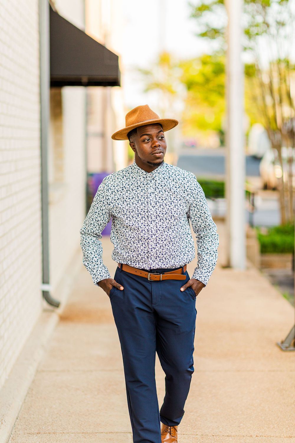 Stylish street portrait featuring brown fedora, patterned shirt and navy pants against white wall.