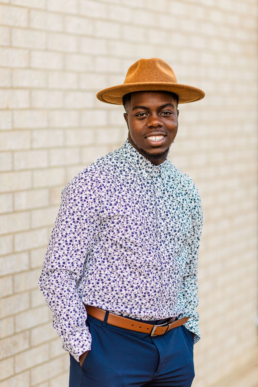 Stylish man in patterned shirt and brown fedora poses against brick wall.