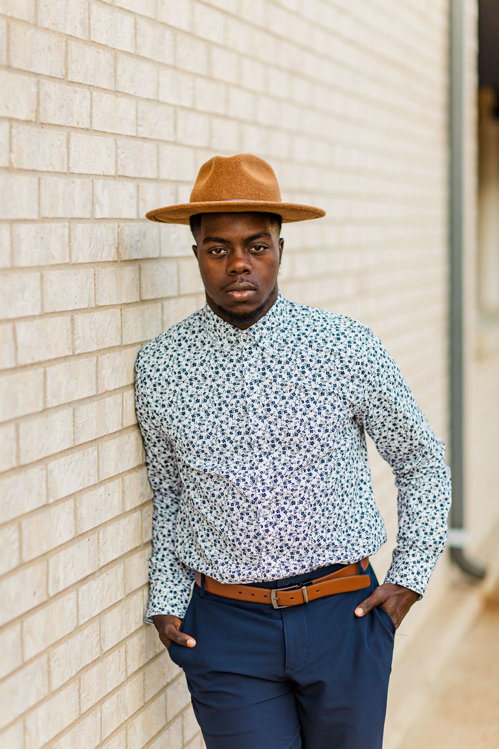 Fashion portrait showing brown fedora and floral print shirt against light brick wall background.