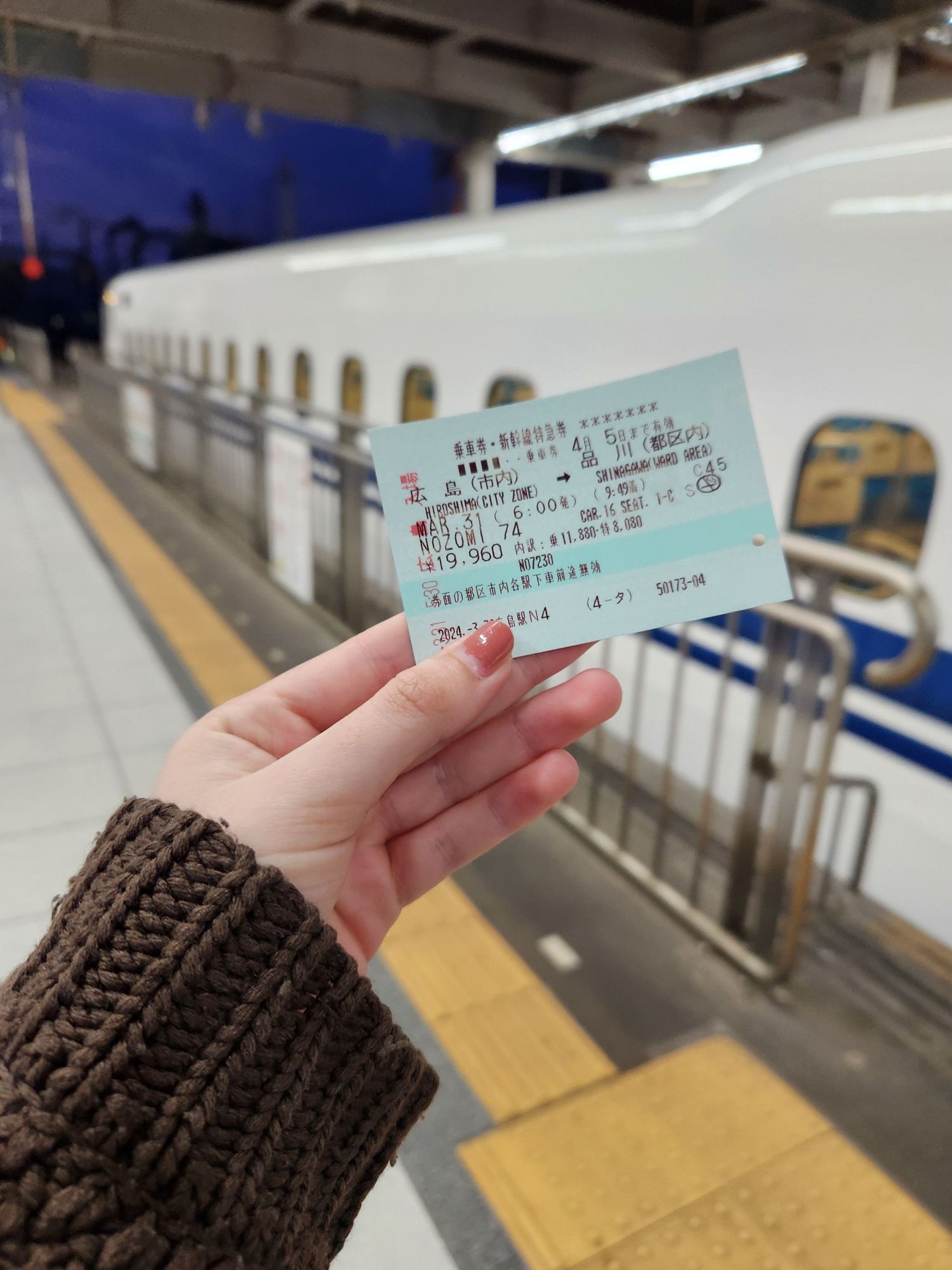 Hand holding a Japanese train ticket in front of a bullet train platform.