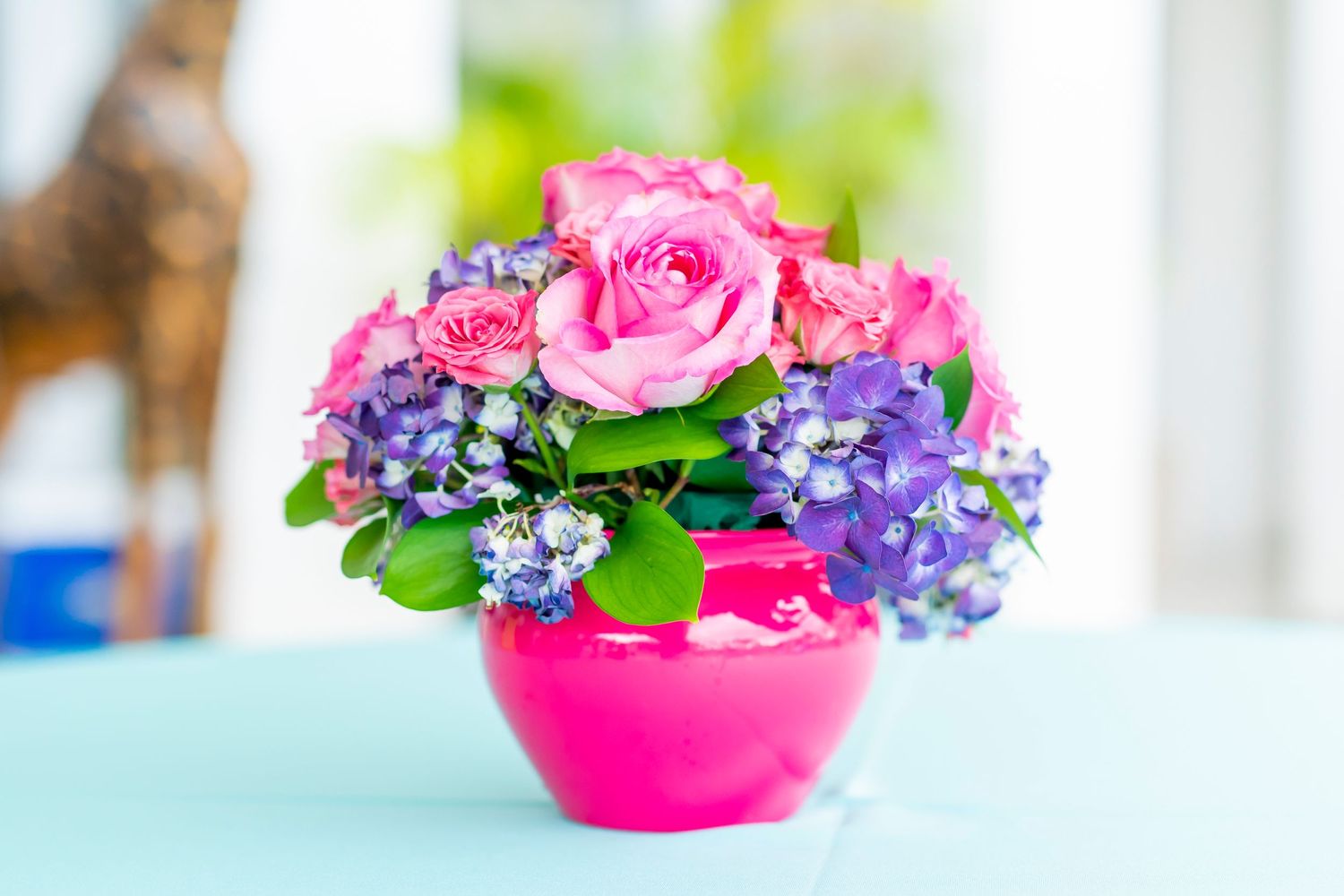 Pink roses and purple flowers arranged in a bright pink vase on a turquoise surface create a cheerful centerpiece.