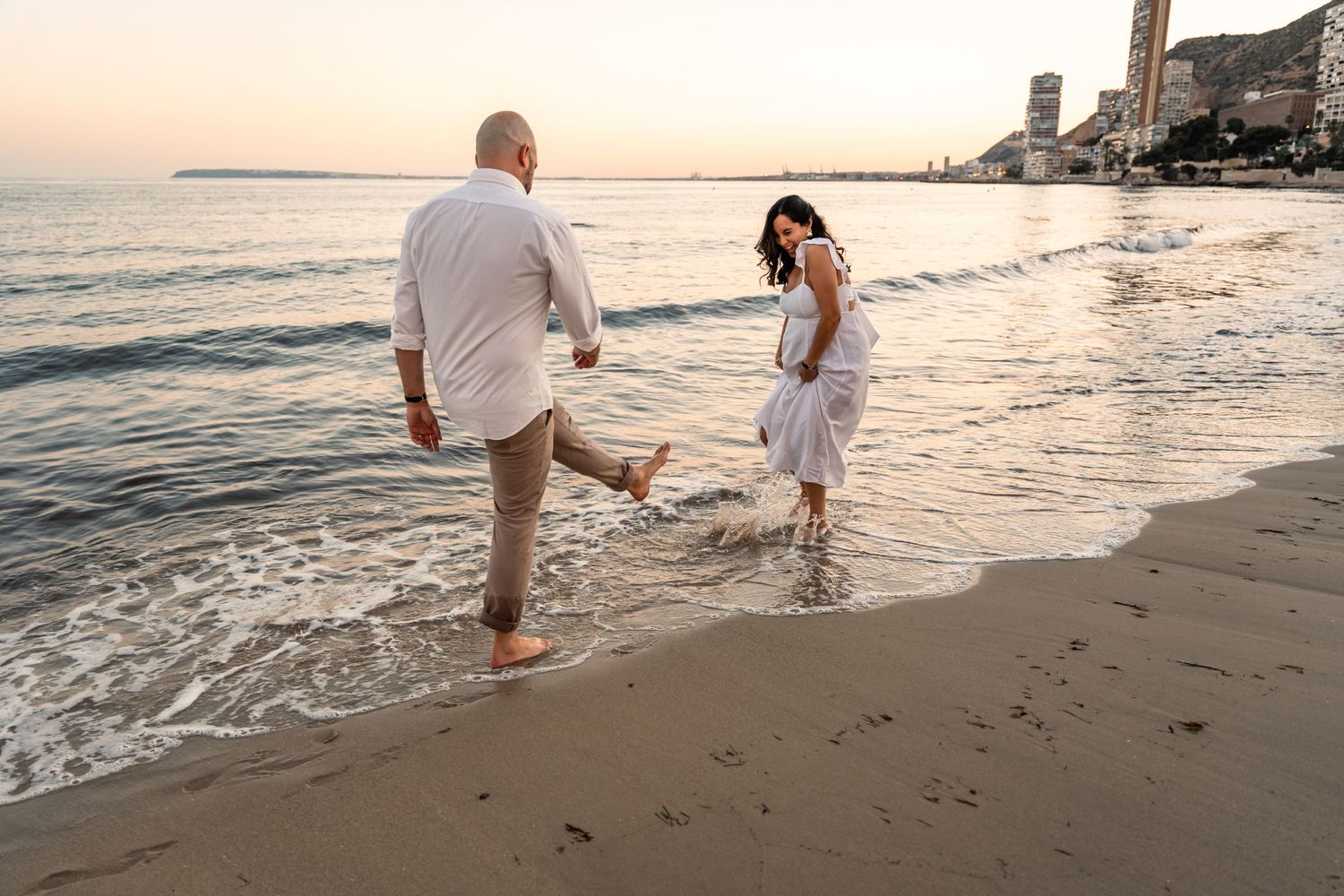 Couple laughing and playfully splashing each other at the beach at sunset wearing white and neutral clothing