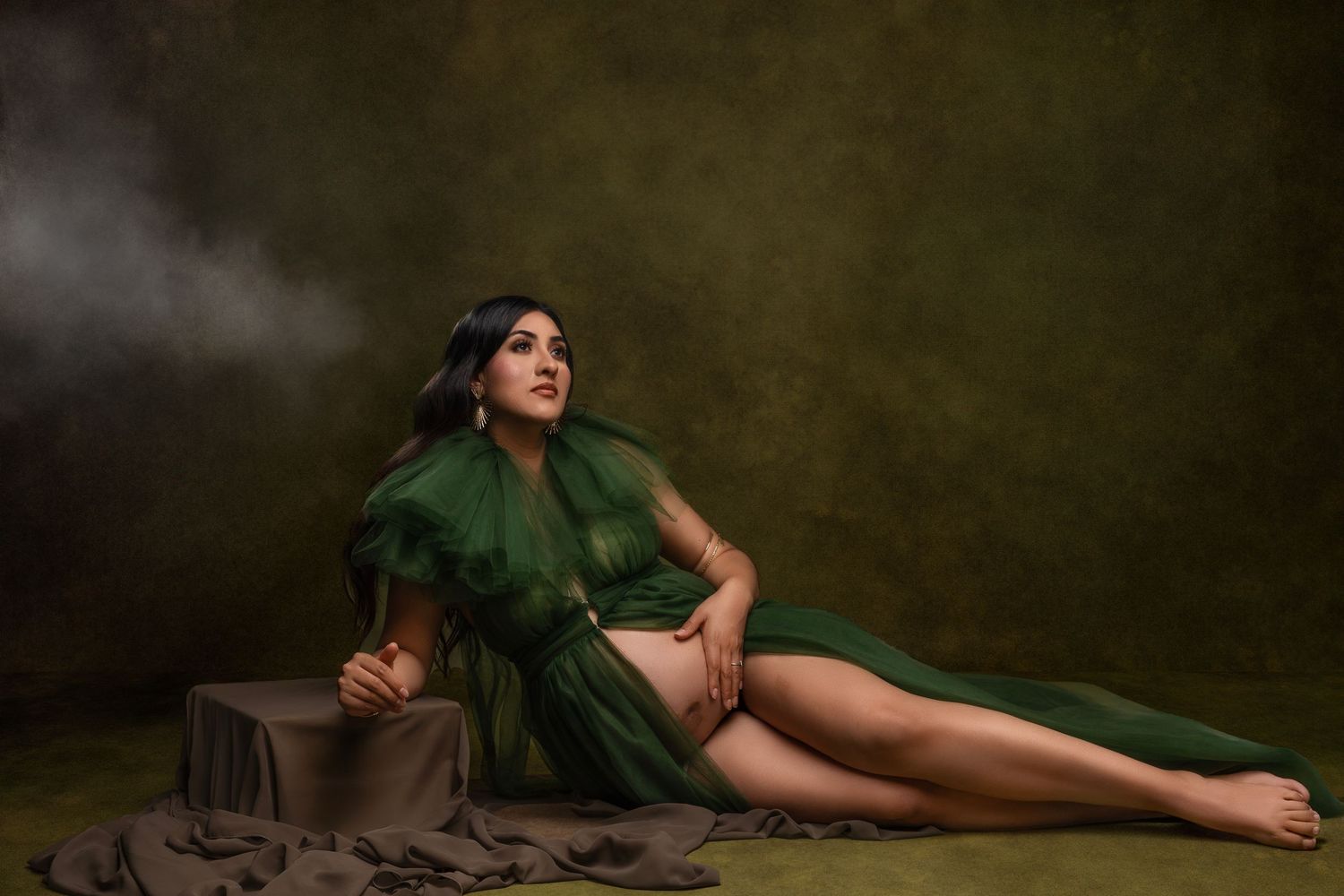 Artistic maternity photo of a woman in flowing green dress reclining on wooden crates in moody studio setting.