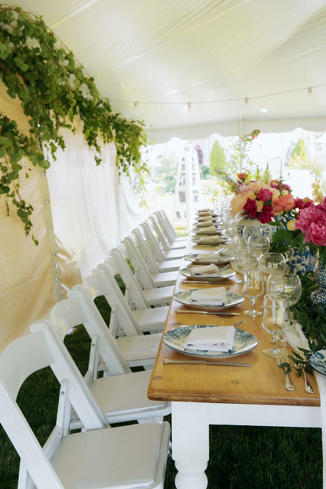 Long rustic wooden table with white chairs set for elegant outdoor dining under tent with greenery.