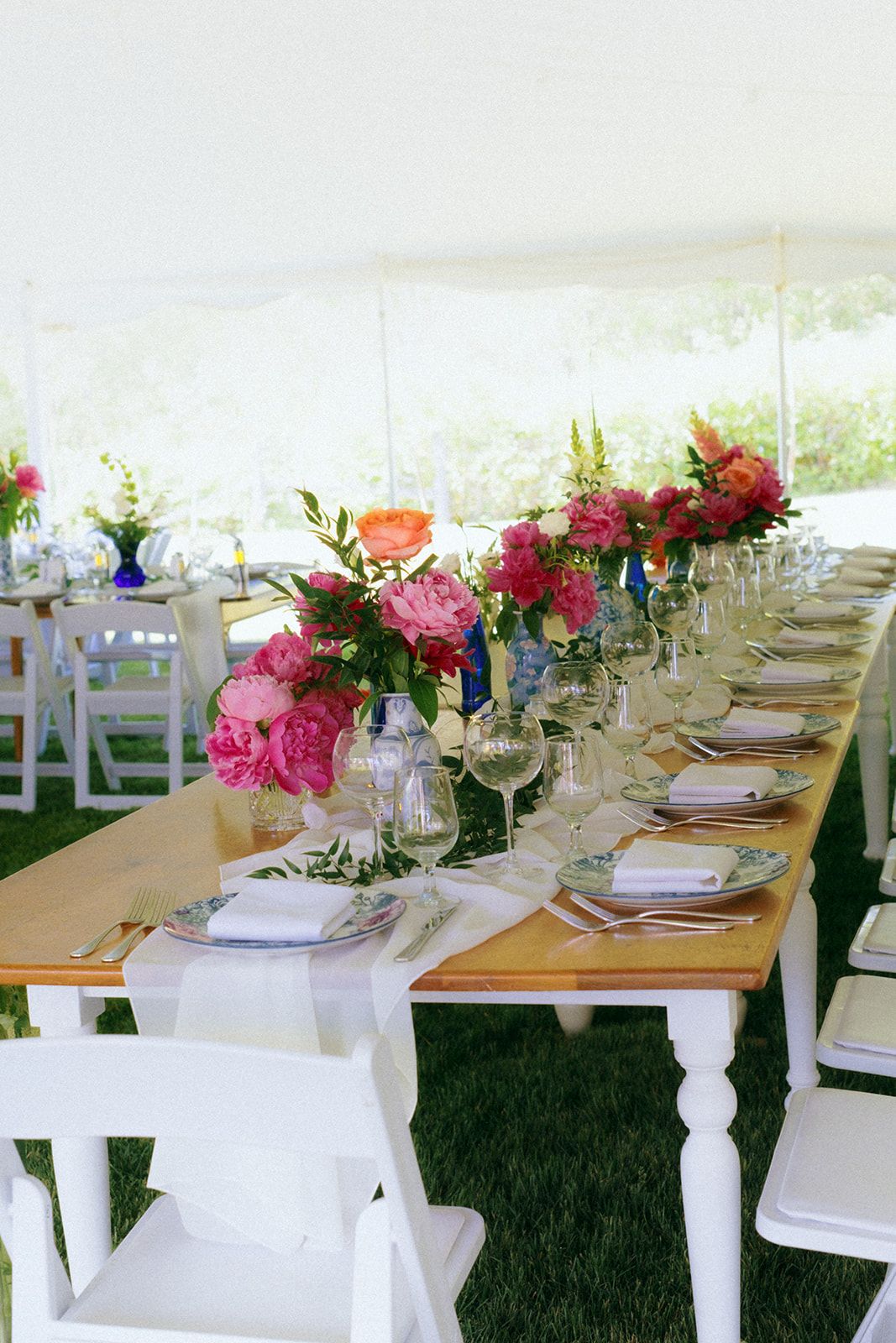 Long wooden farm tables decorated with pink floral arrangements and white place settings under a tent.