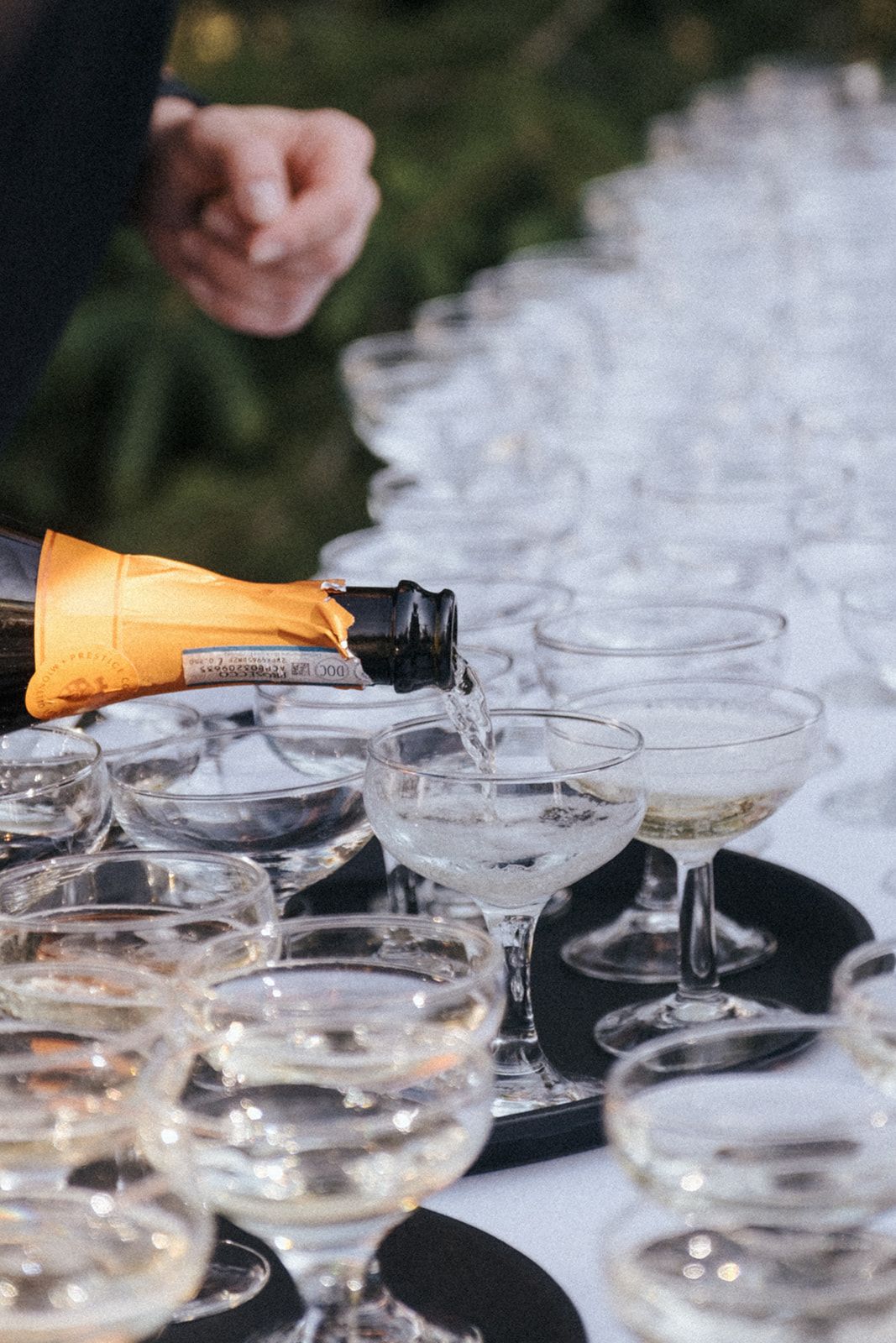 Champagne being poured into rows of coupe glasses on dark serving trays.