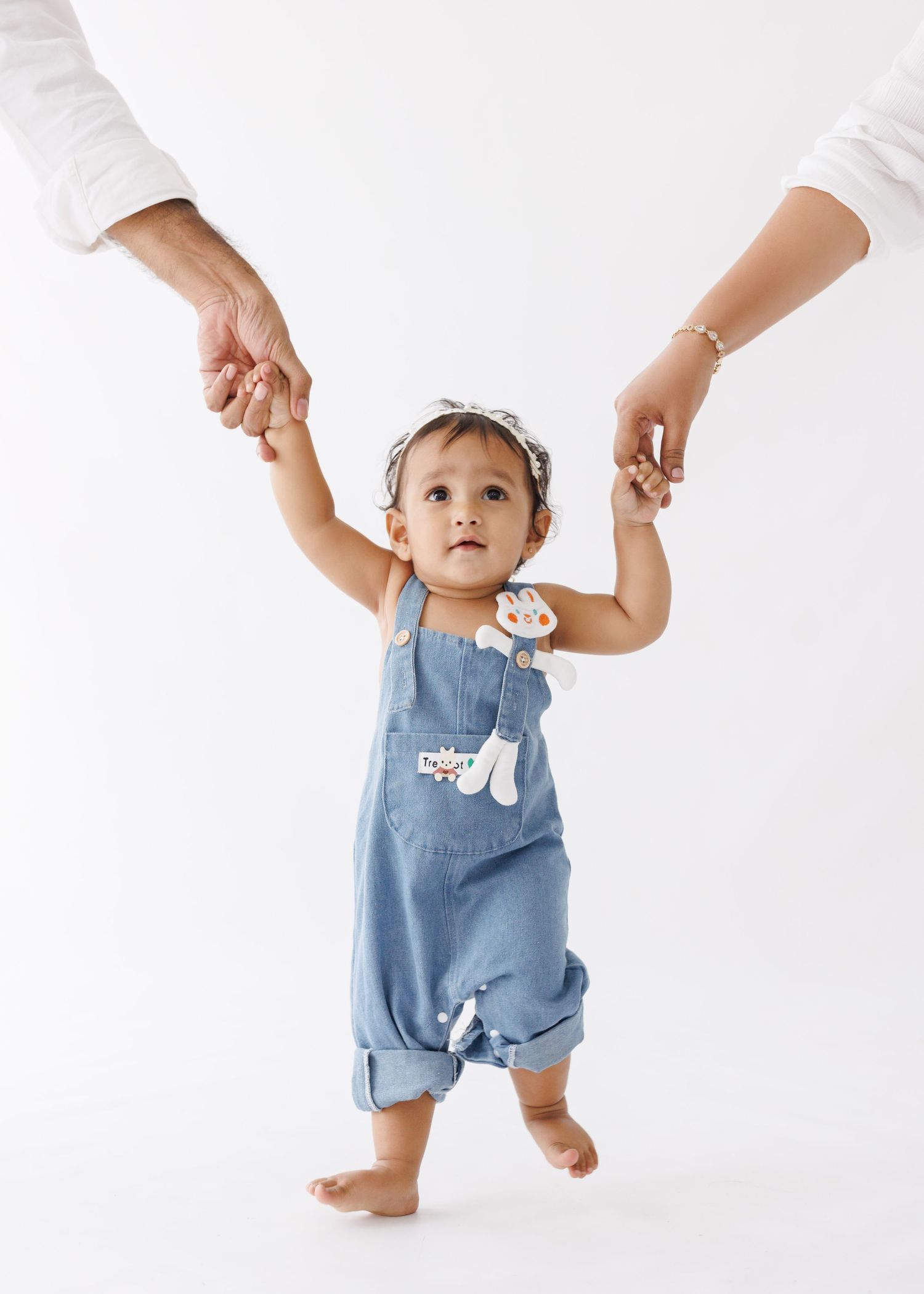 A toddler in denim overalls walking while holding adult hands against white background.