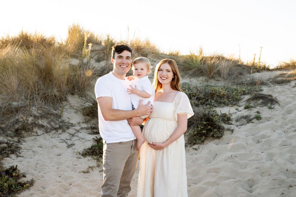 Family wearing white and neutral colors poses together on sandy beach during golden hour sunset.