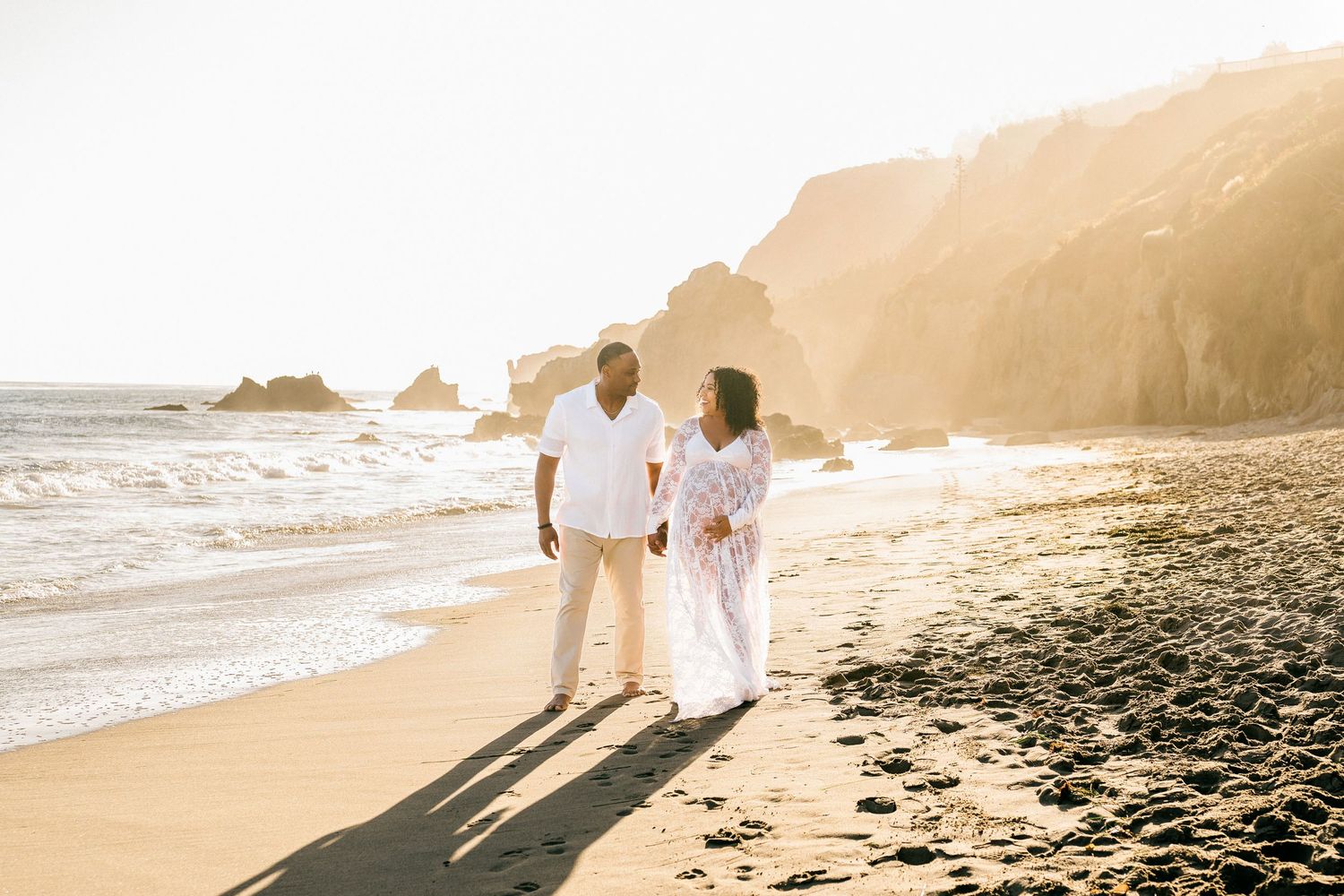 A couple in white clothing walks along a scenic beach at sunset, casting long shadows on the sand.