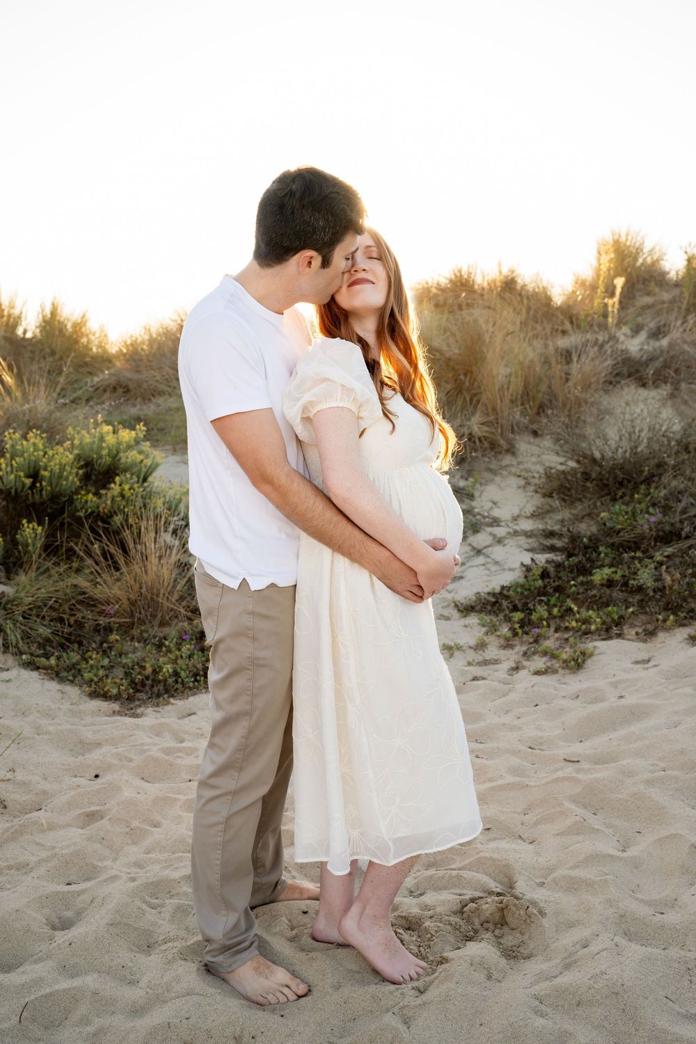 A couple in white and beige clothing share an intimate moment on a sandy beach at sunset.
