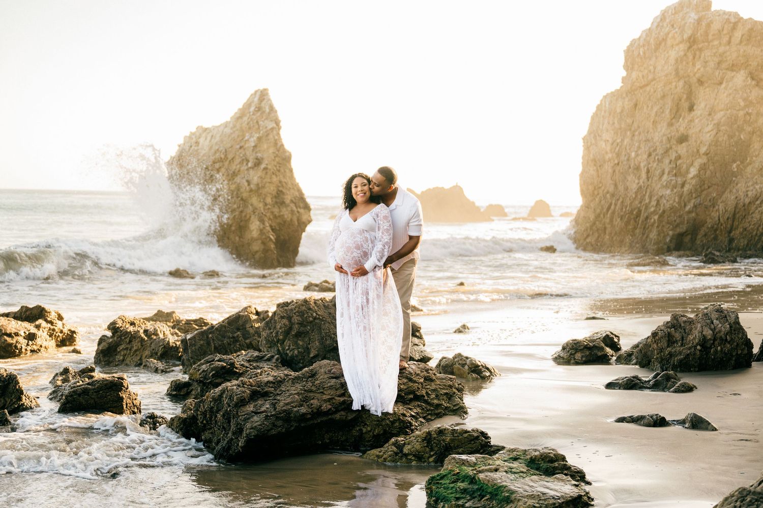 A couple in elegant Maternity attire poses romantically on a scenic beach with dramatic rock formations and ocean waves.