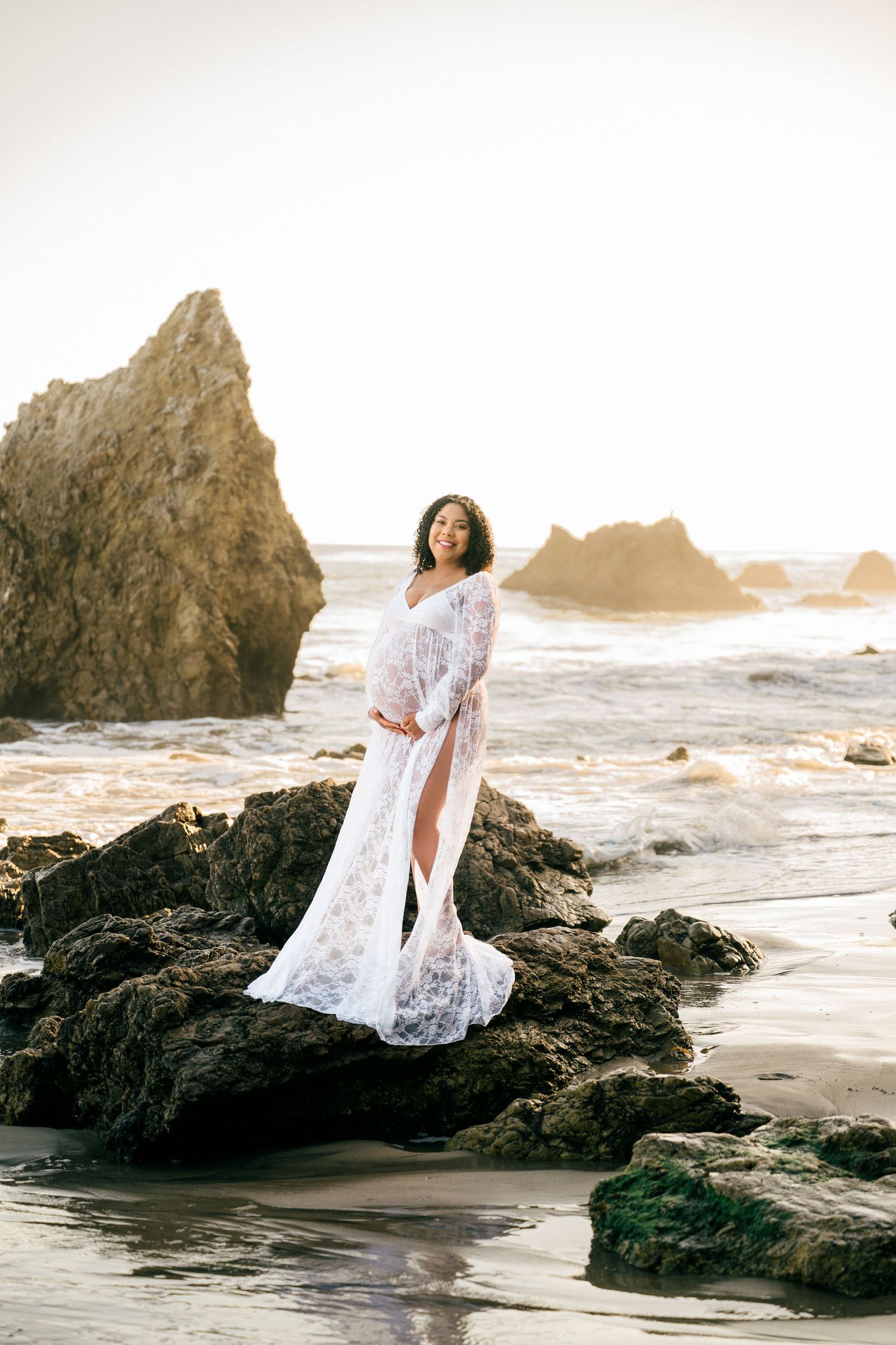 A figure in a flowing white lace dress stands gracefully on rocky coastal cliffs as waves crash against the shore at sunset.