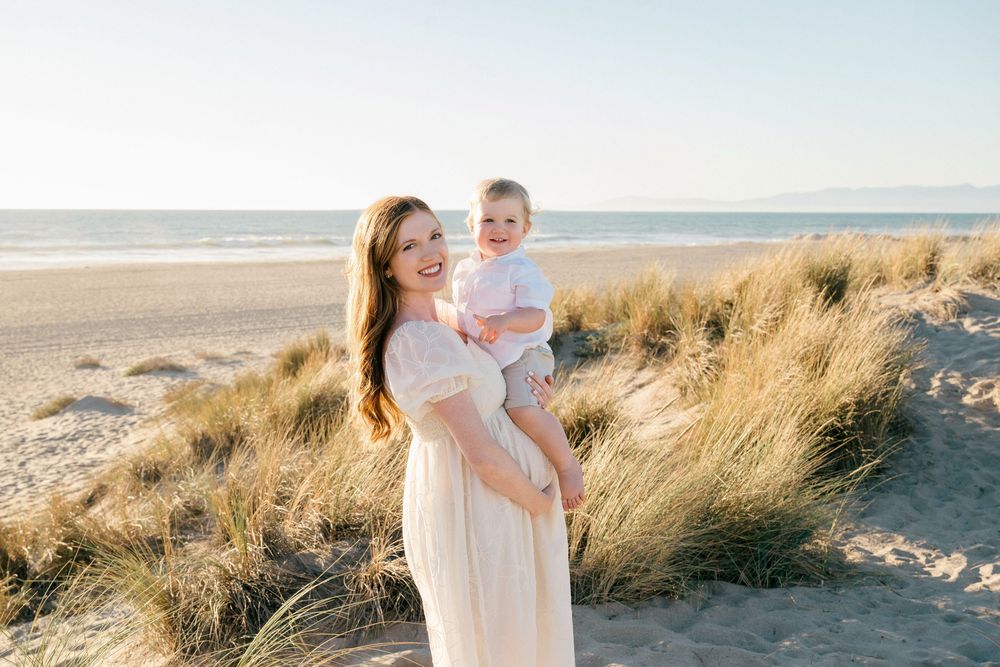 A mother and child share tender moments on a beach with tall grass and dunes during golden hour.