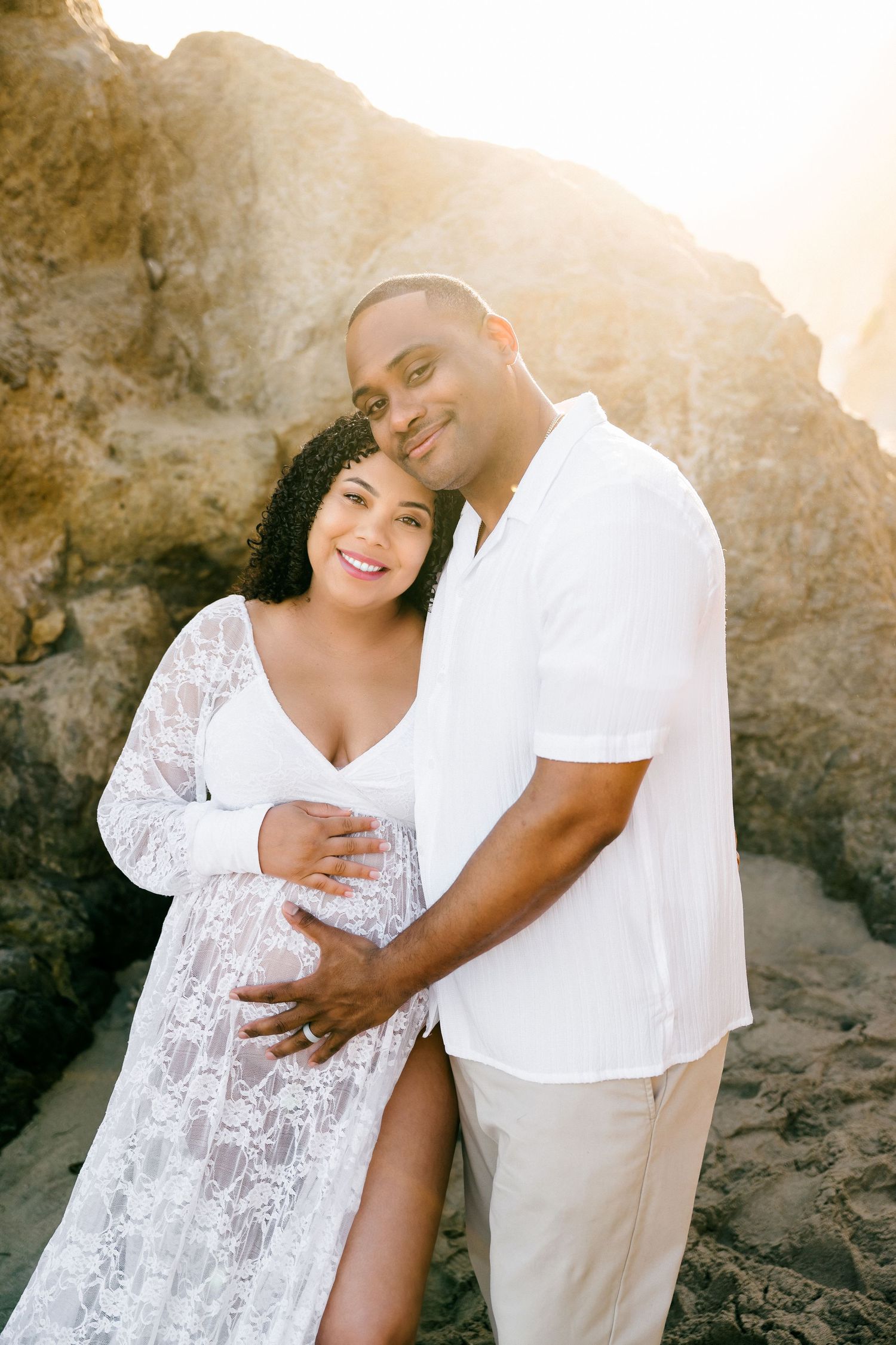 A couple embraces in front of rocky cliffs at sunset wearing white and beige outfits during their maternity photo session.
