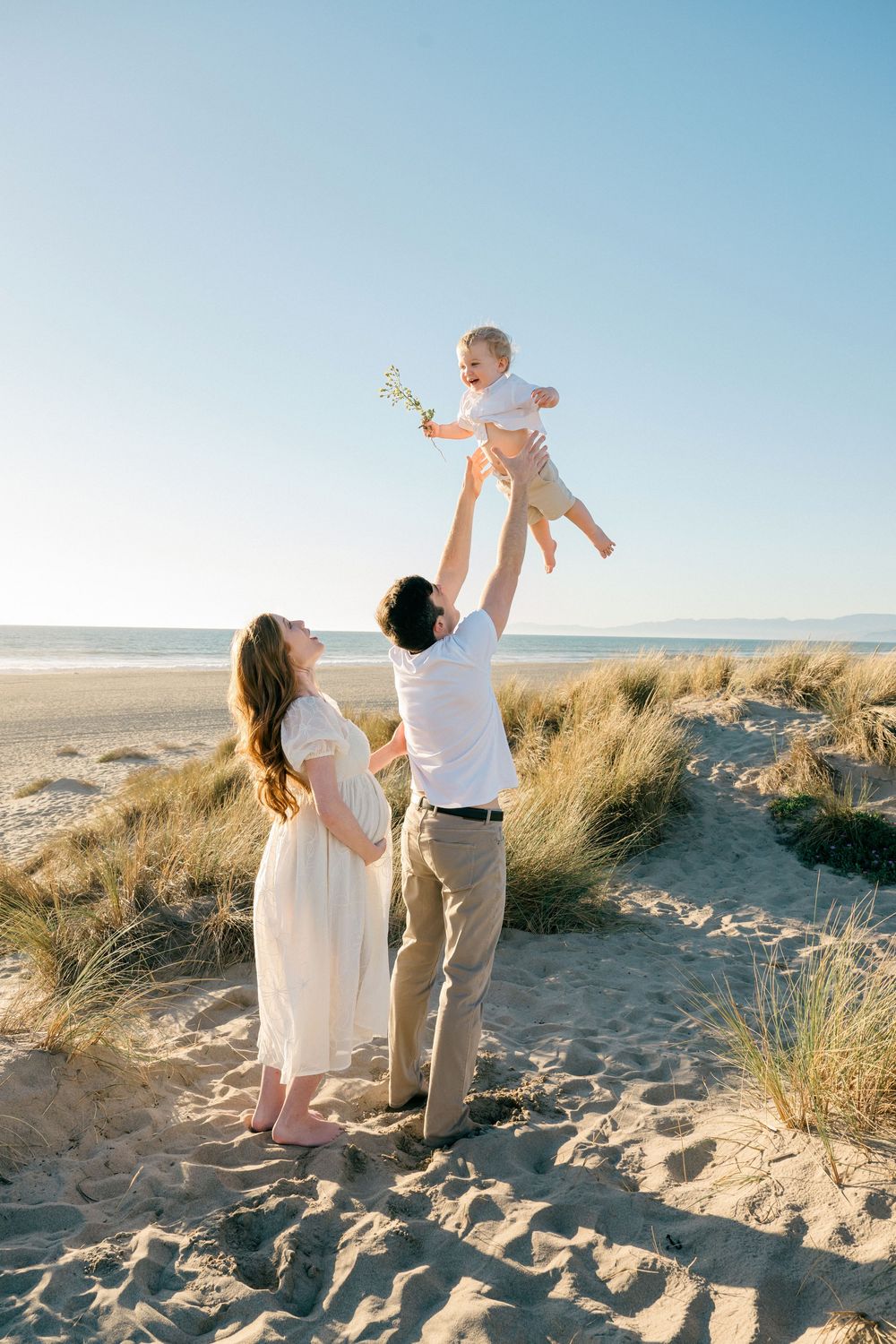 Family enjoying a joyful moment together on a sunny beach with sand dunes and ocean in the background.
