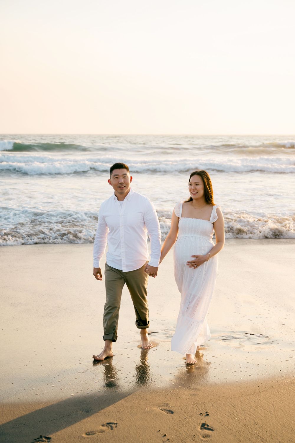 A couple walks hand in hand along a sandy beach at sunset, leaving footprints in the wet sand.