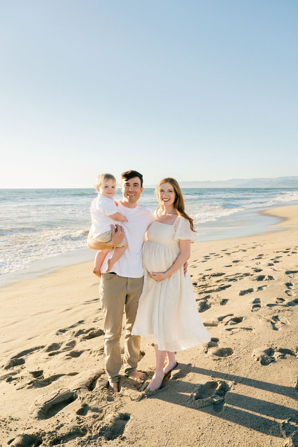 A family walks together on a sunny beach with ocean waves and footprints in the golden sand at sunset.