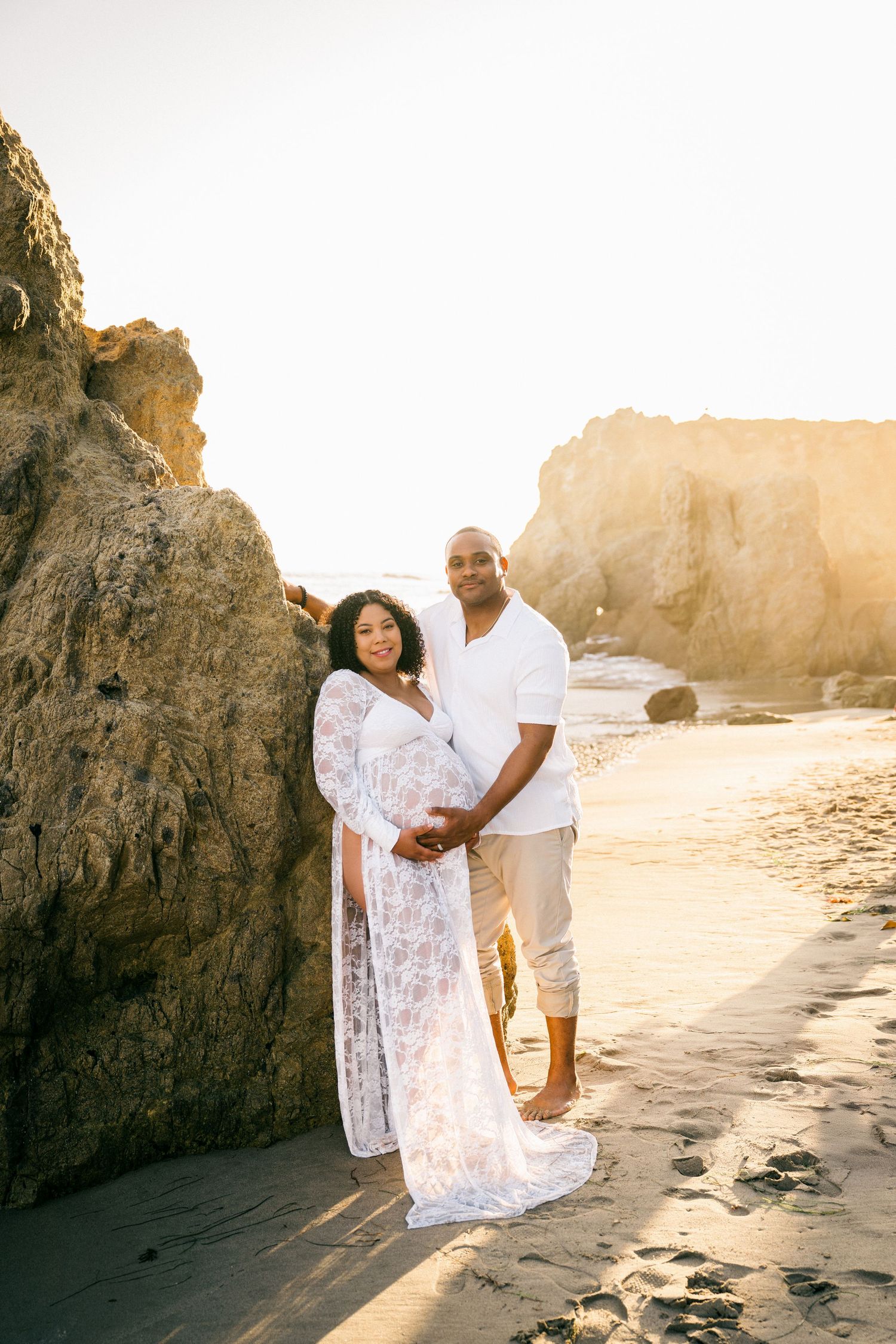 A couple in white clothing shares romantic moments on a sunny beach with rocky cliffs during golden hour.