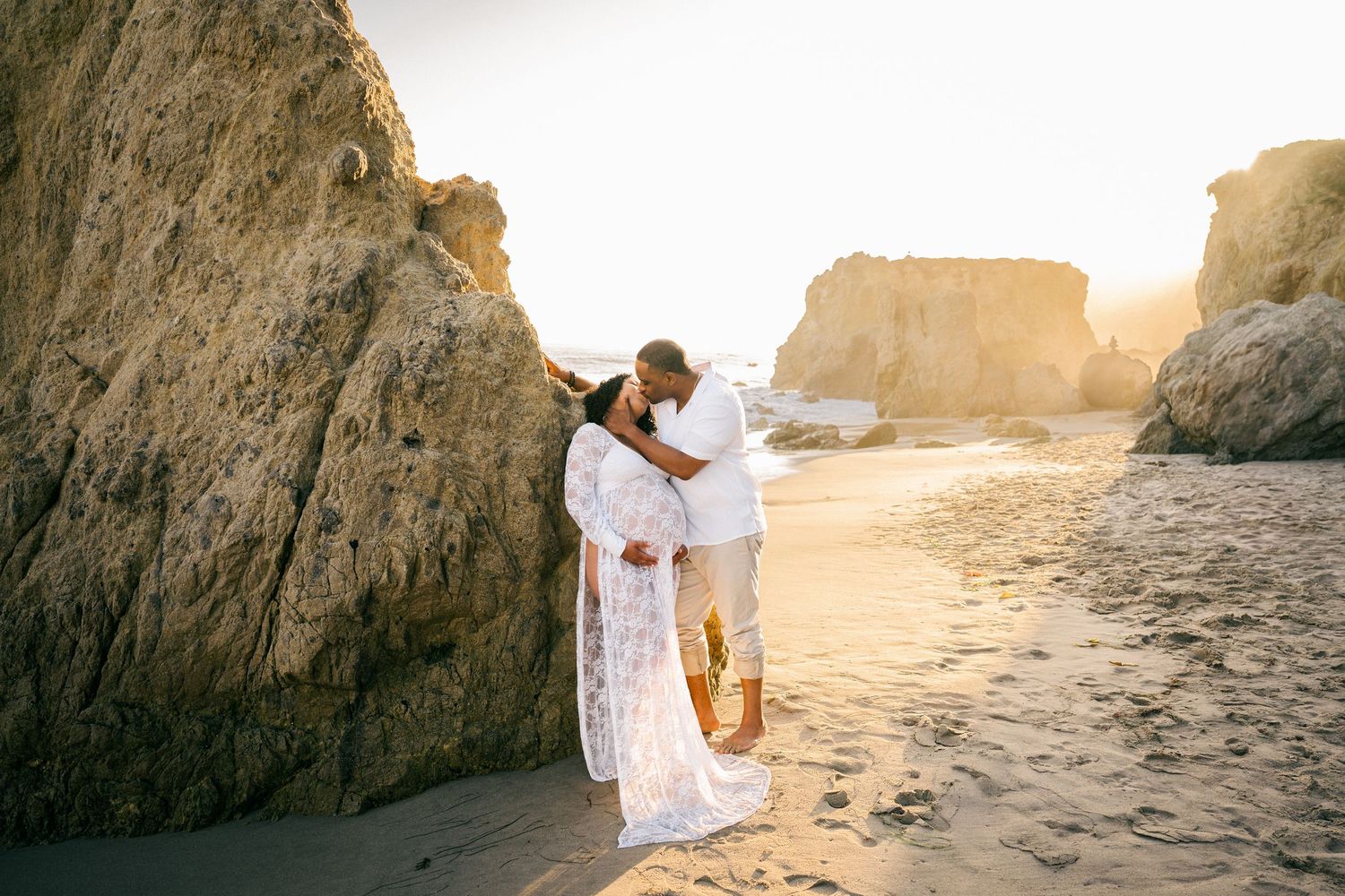 A couple in white attire embracing on a sandy beach at sunset with dramatic rock formations in the background.