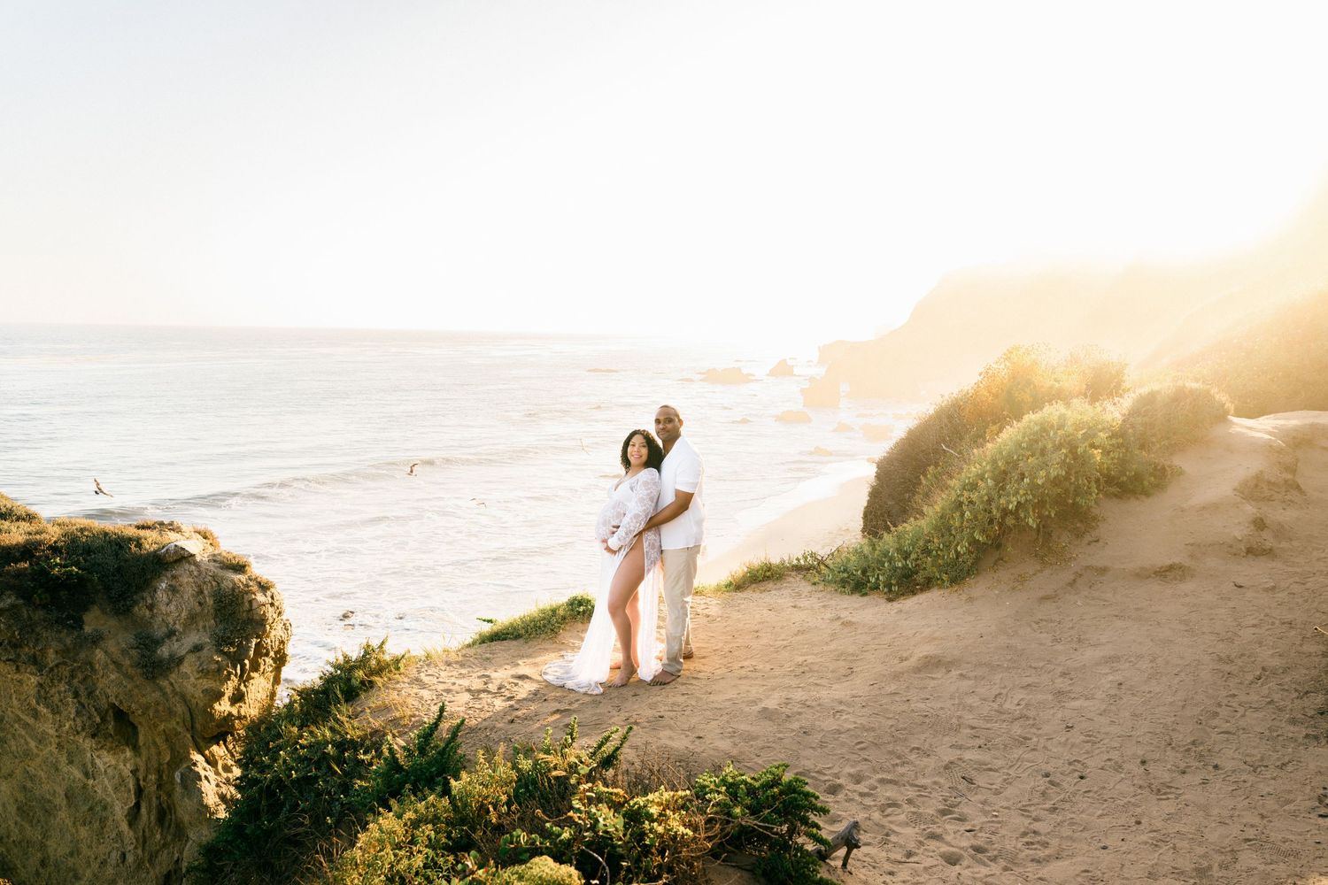 Two people stand silhouetted on a scenic coastal cliff during golden hour sunset.