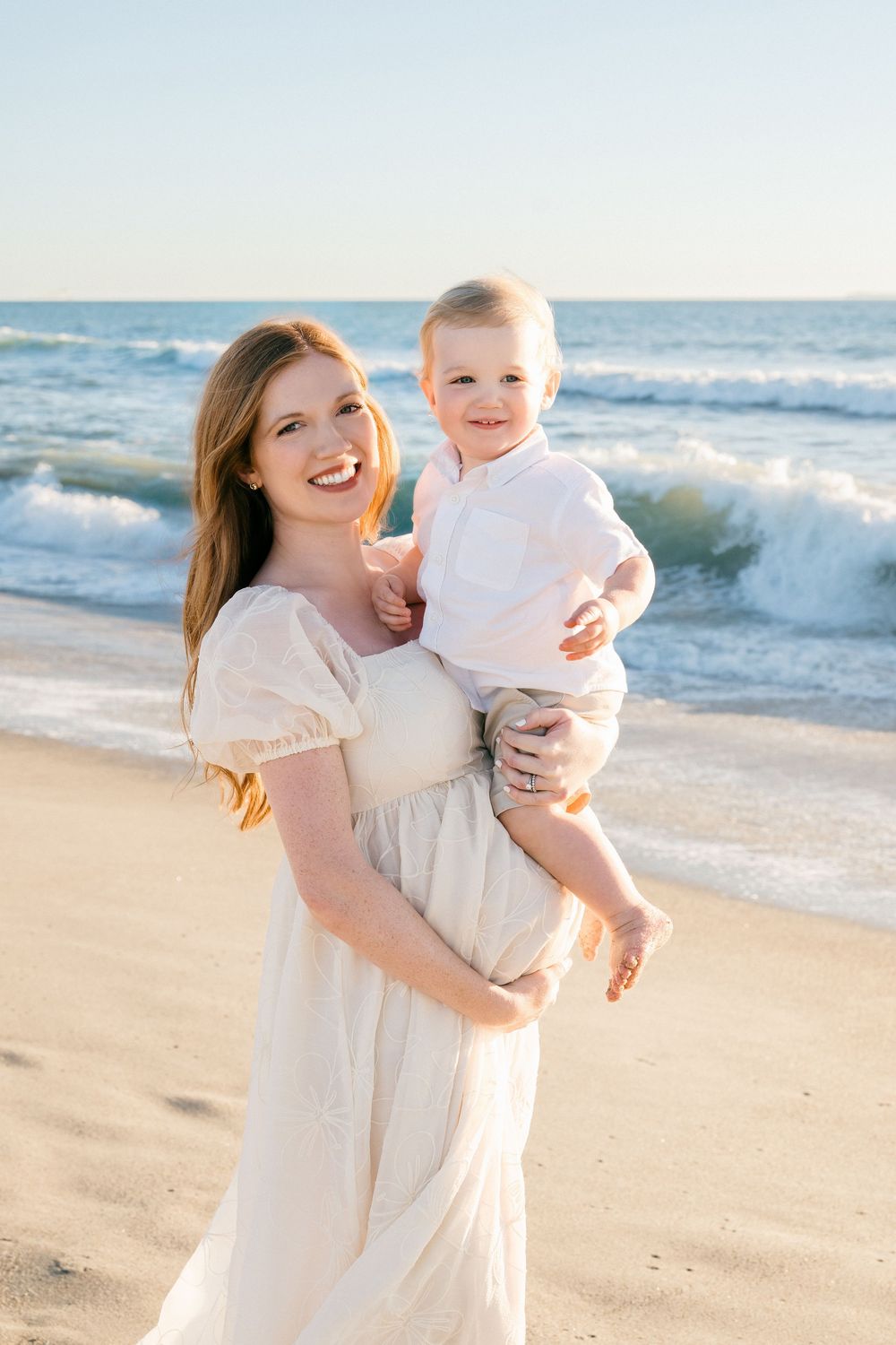 A tender beach photoshoot featuring white dresses against the ocean waves and sandy shore at sunset.