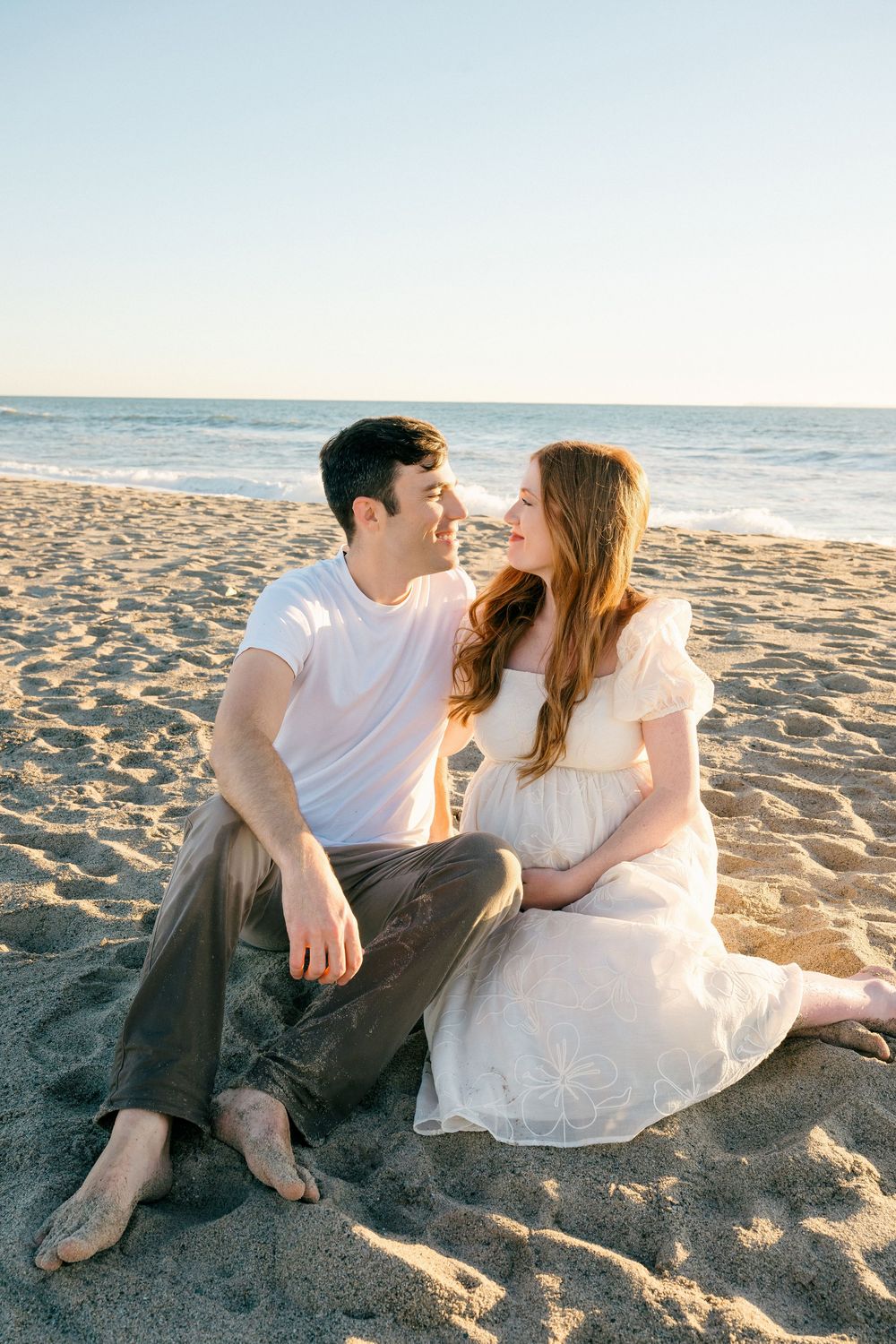 A couple sits together on a sandy beach at sunset sharing an intimate moment by the ocean.