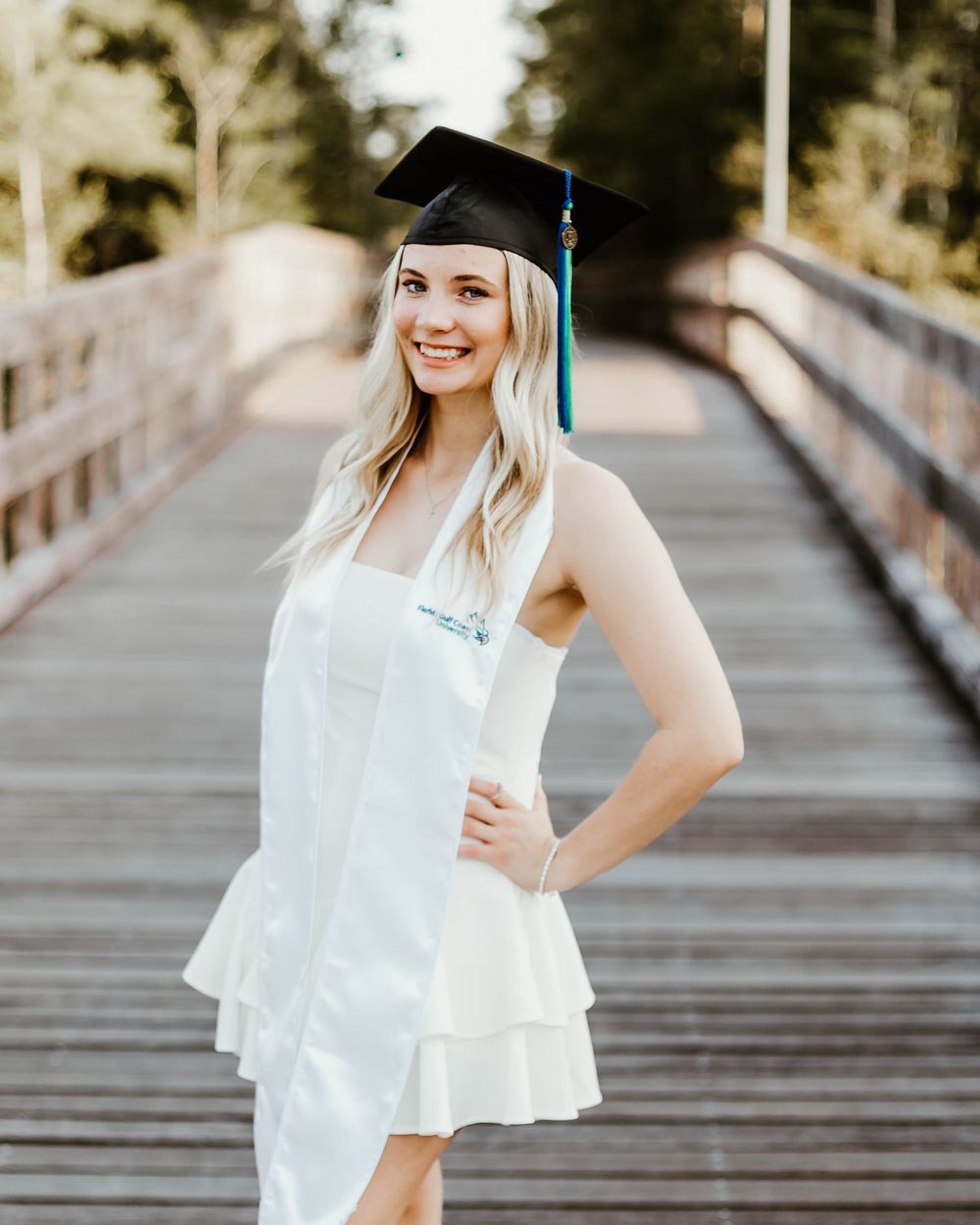 Graduate in white ruffled top poses on wooden bridge wearing graduation cap.
