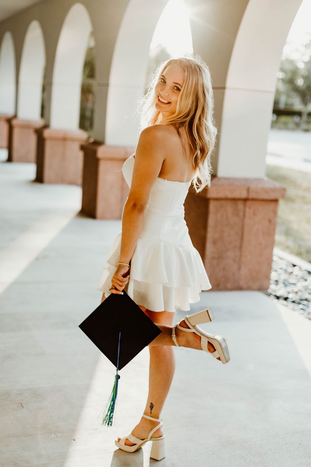 Graduate in white ruffle top celebrates with cap and tassel under archway.