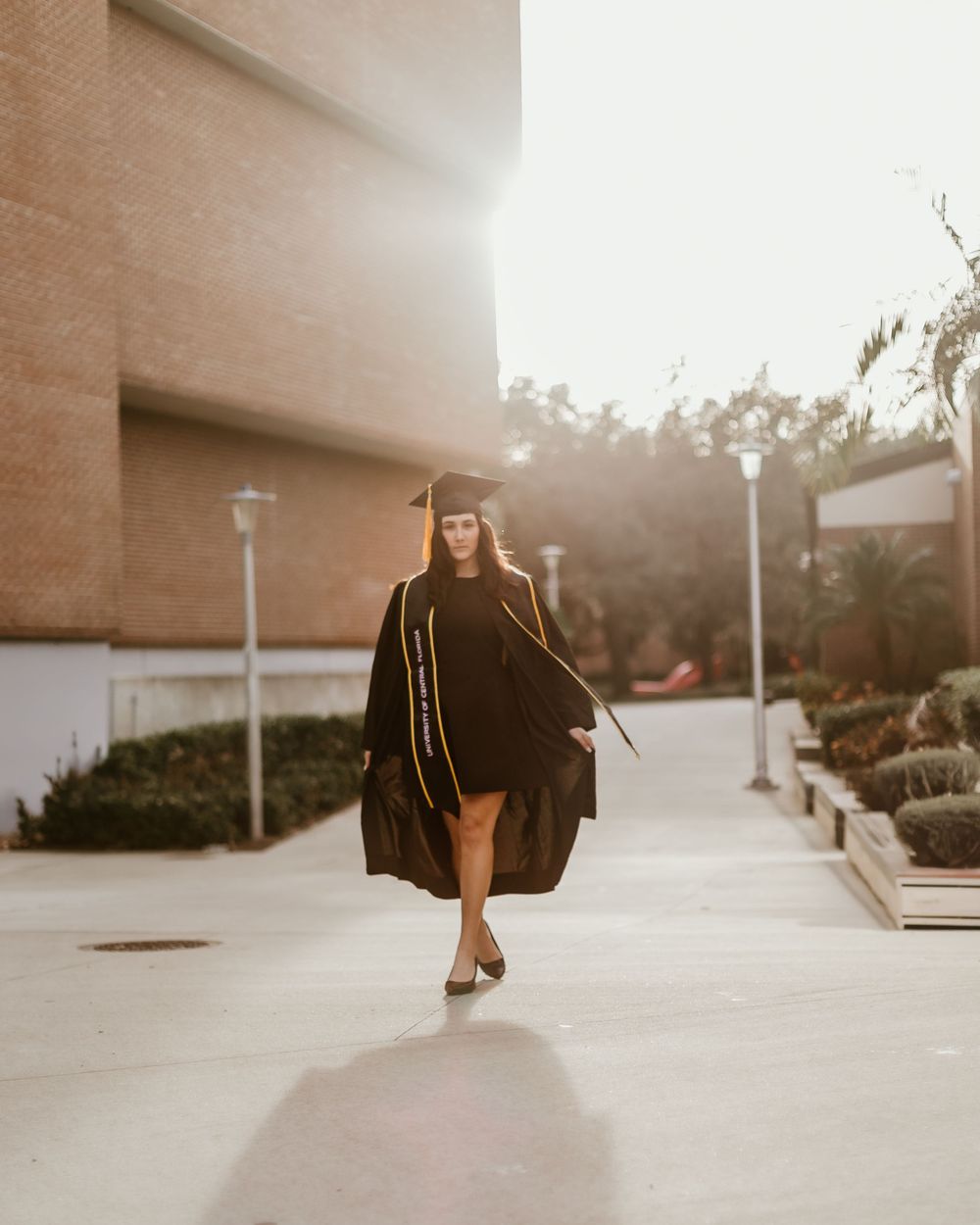 Stylish graduate walks along outdoor pathway at sunset in graduation attire.