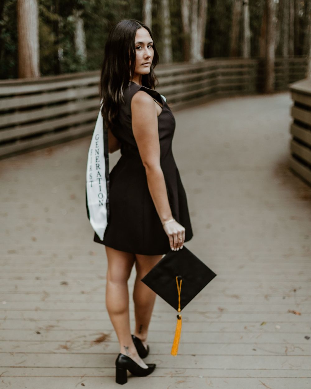 Graduate holding cap and tassel on wooden boardwalk in forest setting.